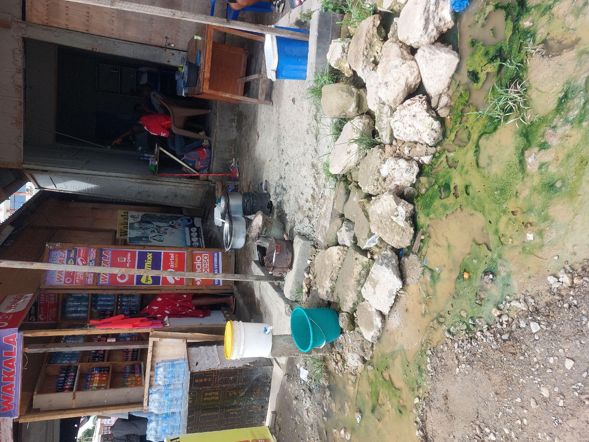 This image captures an outdoor commercial scene in Dar es-Salaam, Tanzania, centered around a small, open-fronted shop or kiosk. The structure, appearing to be made of wood and plywood, functions as a "WAKALA" (agent) for various mobile money services. Numerous vibrant signs adorn the shopfront, featuring brands such as "m-pesa," "airtel Money," "halopesa," "yasmixx," "VODA LIPA," and "airtel."
Inside the shop, shelves are stocked with bottled drinks, including water and sodas. A person in a red shirt is visible, seated and attending to the business, with another individual partially obscured in the darker interior. A white bucket with a tap, possibly for handwashing or water dispensing, sits near a green bucket on the ground. Various metal pots and a blue cooler are also visible.
Adjacent to the shop, the ground is unpaved and uneven, leading to a prominent muddy ditch filled with stagnant, green-algae-covered water. A pile of large rocks borders this water, suggesting an informal drainage system. The scene is illuminated by natural daylight, indicating a bright, sunny day. Visible text also includes "Marine Board" and "大众木业" (Public Wood Industry) on wooden crates. The overall impression is of daily commerce in a bustling, semi-urban environment, characterized by improvised infrastructure and visible environmental challenges related to drainage.