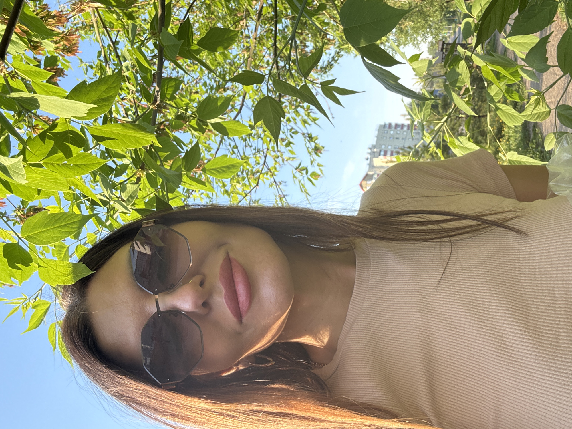 The image depicts a woman wearing sunglasses, framed by green foliage. The background reveals a cityscape, with a multi-story apartment building visible. Based on the details of the buildings and vegetation, this image was taken in the city of Chaikovsky, Russia. The woman is wearing a light-colored shirt. The image is taken from a low angle, looking upwards.