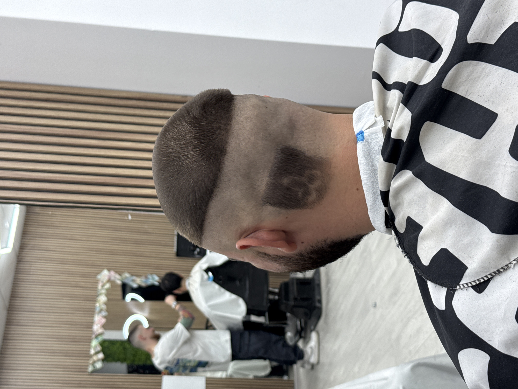 A barbershop interior in Sofia, Bulgaria, features a male customer in the foreground, viewed from behind, wearing a black and white patterned barber's cape over a white neck strip. His dark hair is styled with a high skin fade, a distinct horizontal separation line for the longer top section, and a rectangular design shaved into the back of his head. His right ear and a dark beard are visible.
Reflected in a large wall mirror in the midground, a male barber, with short light hair and visible arm tattoos, wearing a graphic white t-shirt and dark trousers, is actively cutting the hair of a second male customer. This customer is seated in a black barber chair, draped in a white cape, facing away from the mirror.
The barbershop environment includes a light-colored vertical wooden slat wall visible in the reflection, a light-toned floor, and a white ceiling with linear lighting. The mirror itself is adorned with paper notes and artificial green foliage along its top edge, alongside two circular light fixtures. A plain white wall is present to the right of the foreground subject.