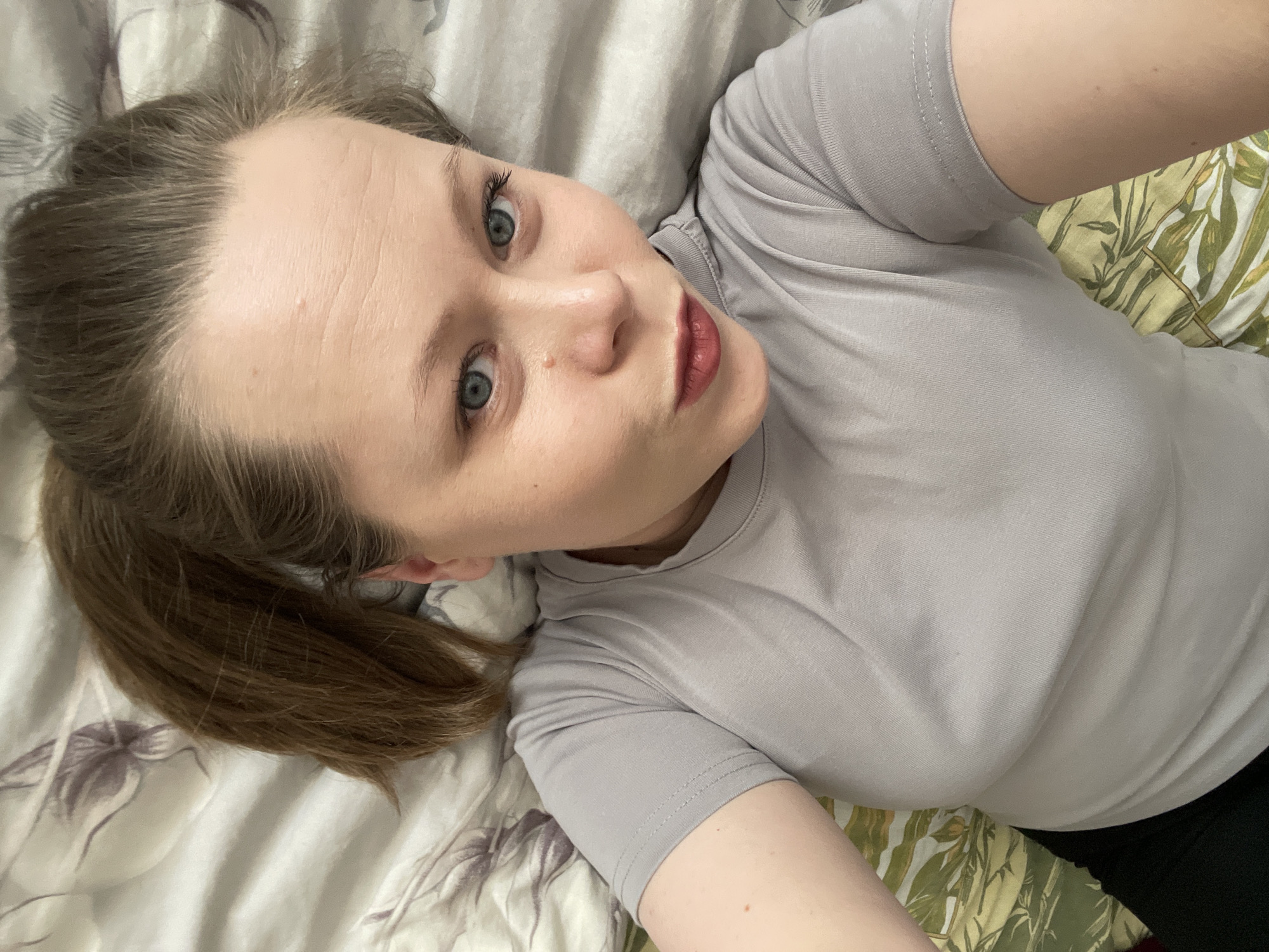 A young adult female is depicted in a close-up, high-angle selfie, lying down on what appears to be a bed in Chaikovsky, Russia. Her head is slightly tilted, and she is looking directly into the camera with wide, striking blue-green eyes. She has light brown hair, pulled back into a low ponytail, and fair skin with faint lines visible on her forehead. A small mole is noticeable on the bridge of her nose. Her lips are adorned with a reddish-pink lipstick and are slightly puckered, giving her a subtle pout.
She is wearing a simple light gray short-sleeved t-shirt. The background consists of two different patterned fabrics, likely bed sheets or pillowcases. One pattern features muted gray and purple floral or leaf designs, while the other is a more vibrant green and yellowish-green botanical print. The lighting is soft and even, typical of an indoor setting. There is no visible text in the image. The overall mood is casual and personal.