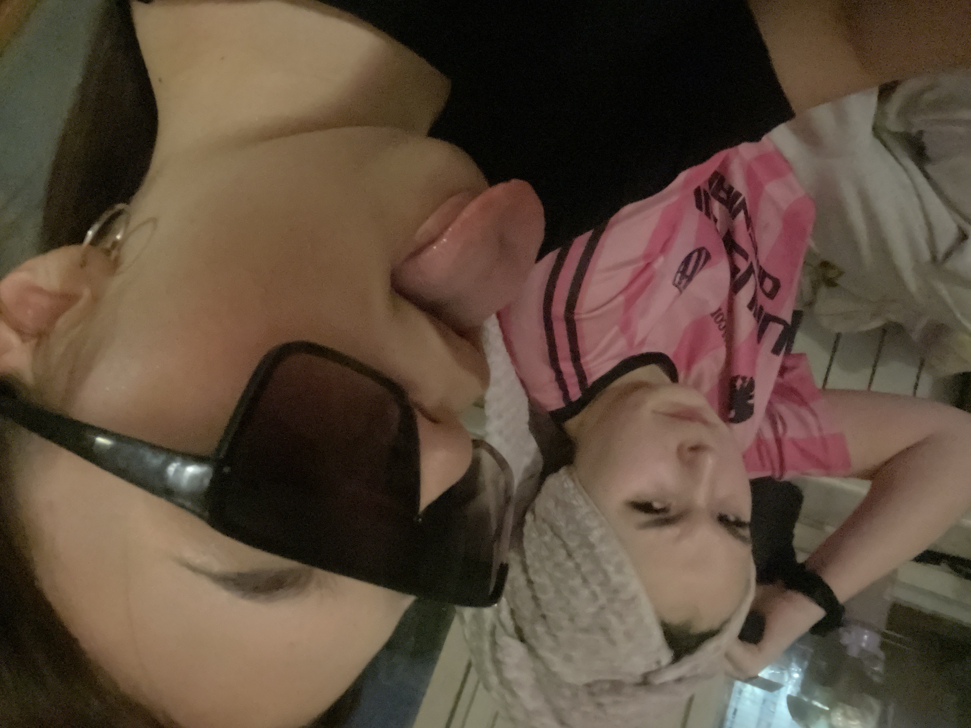 This close-up photograph features two young individuals posing in an indoor setting. The image is taken from a slightly elevated perspective.
The person on the left foreground has fair skin and dark hair, and is wearing dark sunglasses that cover their eyes. A silver hoop earring is visible in their left ear. They are making a playful expression by extending their tongue from their mouth. A dark garment is visible on their upper body.
The second person, positioned slightly behind and to the right, also has fair skin and is looking directly towards the camera with a neutral expression. They are wearing a pink short-sleeved top adorned with black vertical stripes and the word 'ADIDAS' printed in black text along one of the stripes. A light grey or beige knitted headband or scarf is worn around their head. A black band, possibly a hair tie, is visible on their right wrist.
The background is blurred and indistinct, showing subtle reflections, possibly from a table or glass surface. The overall atmosphere is casual and posed. While the context states the location is Murino, Russia, there are no visual cues within the image itself to confirm this.