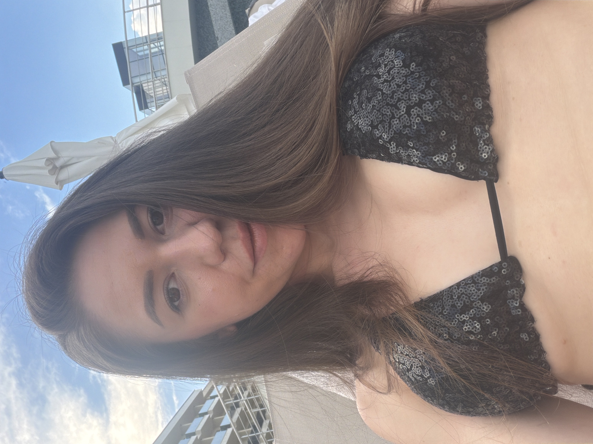 A young woman is lying down outdoors, wearing a black sequined bikini top. Her long brown hair is spread out around her. The background shows a bright blue sky with some clouds, the top of a modern building with geometric windows, and a white umbrella. The scene appears to be a sunny day, likely during daylight hours. The woman is looking towards the camera, and her expression is neutral or slightly contemplative. The overall setting suggests a resort or a rooftop area, possibly in Hisarya, Bulgaria. There is no visible text in the image.