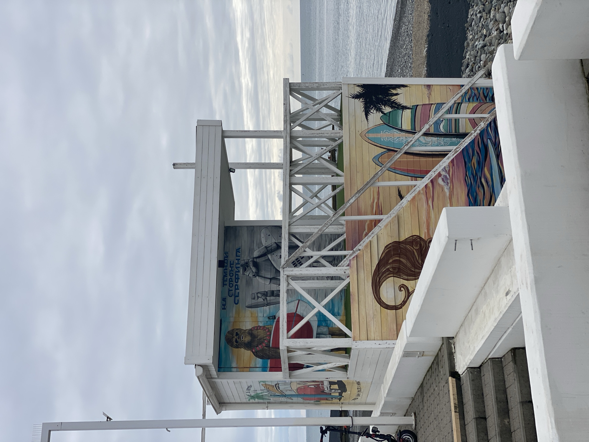 This image depicts a two-story, white-painted wooden beachside structure situated on a pebbled beach adjacent to the Black Sea in Sochi, Russia, under an overcast sky. The structure's exterior is adorned with colorful murals. The upper level features a white wooden railing with an "X" pattern, and a set of wooden steps with a matching railing ascends to this level from the right.
The most prominent mural, on the front wall of the ground level, illustrates Chewbacca from Star Wars holding a red surfboard, standing next to a Stormtrooper, also holding a surfboard. Above these figures, Russian text reads "НА ТЕМНОЙ СТОРОНЕ СЕРФИНГА" (On the Dark Side of Surfing). A partial mural on the left side of the structure shows a red Volkswagen van with a surfboard on its roof, palm trees, and text partially visible as "LIFE IS BETTER...". The lower section of the structure's walls features additional murals of stylized ocean waves, multiple surfboards, a palm tree silhouette, and a large brown swirling graphic.
To the left of the structure, two electric scooters are parked on a grey paved surface next to a tall, white vertical pole. Dark grey concrete steps lead down into the foreground from this paved area, bordered by low, white concrete retaining walls. The sea, characterized by a calm, grey-blue surface, stretches across the right half of the background under a uniformly cloudy sky. The immediate foreground consists of more white concrete structures and the aforementioned steps. No individuals are present in the scene.