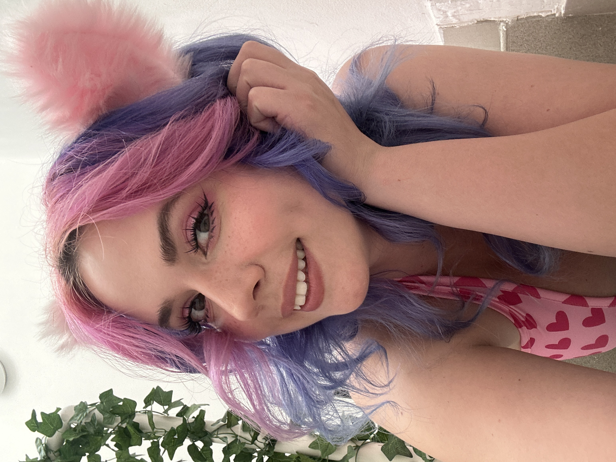 The image shows a woman with pink and purple hair and pink cat ears smiling at the camera. She is wearing a pink top with red hearts. Some greenery is visible at the bottom left of the image, and a wall appears at the top right. The location is not evident from the image.