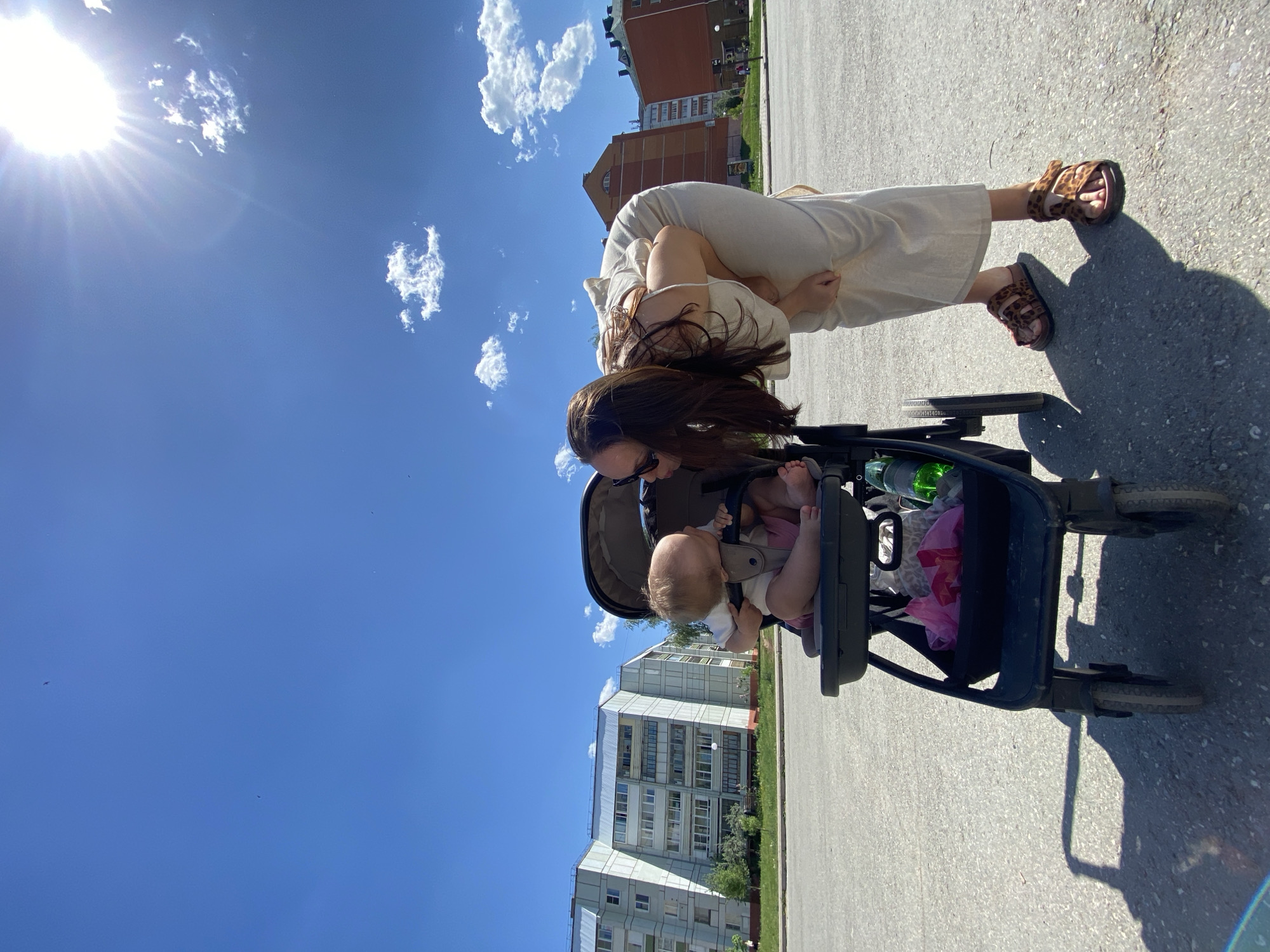 A woman with long brown hair and sunglasses bends over a baby in a black stroller on an asphalt surface, possibly a sidewalk or road. The baby sits in the stroller, and the woman appears to be interacting with the child. The woman wears a light-colored dress and sandals with a leopard print pattern.
In the background, there are buildings that appear to be residential apartment blocks. The style and architecture, along with information available in the metadata, indicates the location is Chaikovsky, Perm Krai, Russia. The sky is blue with scattered white clouds, and the sun is shining brightly, casting shadows of the woman and stroller on the ground. The scene suggests a sunny day in an urban environment.