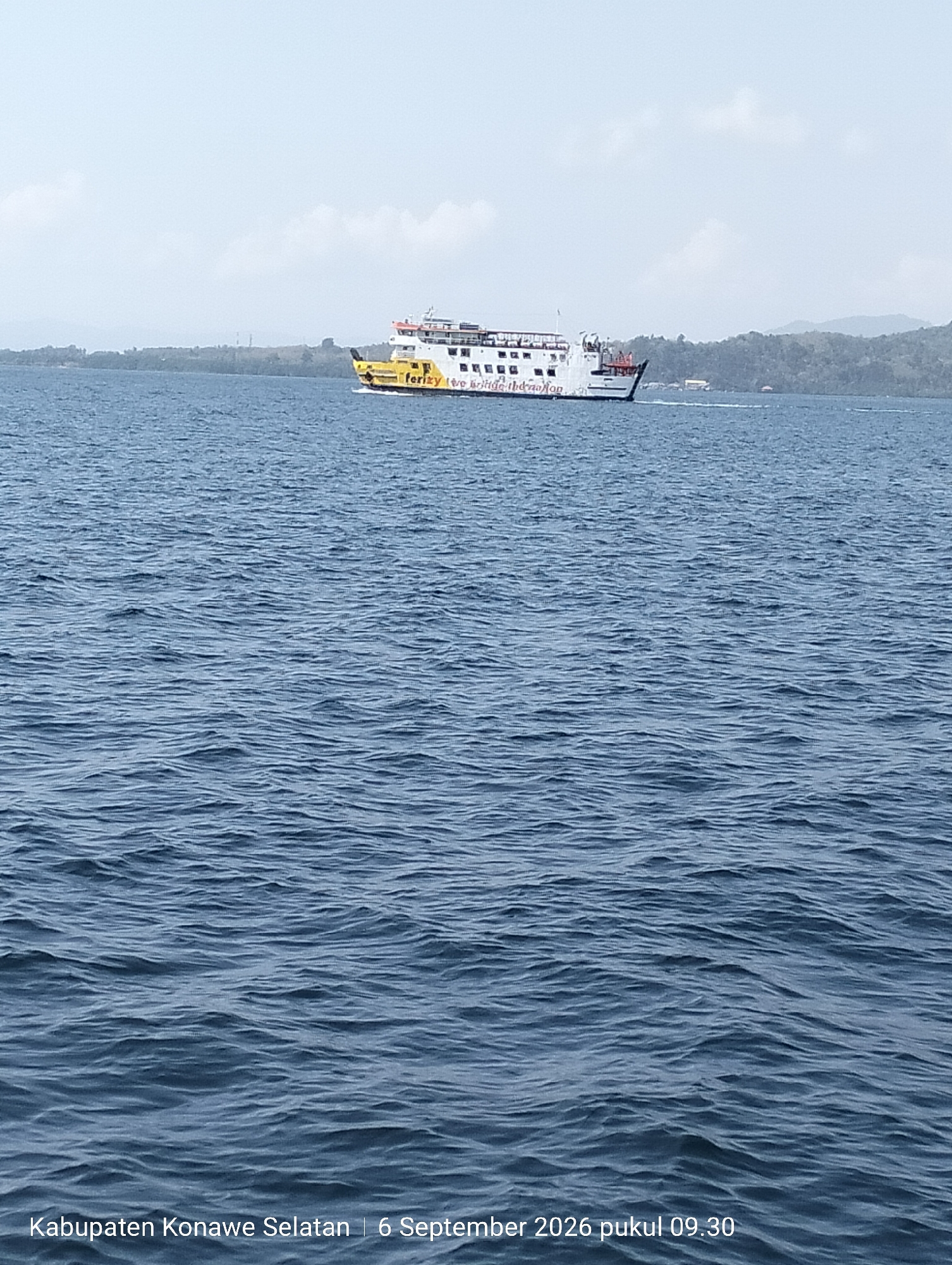 A ferry boat travels across a large body of water under a hazy sky. The ferry is white with a yellow stripe and red accents, and it has the words "ferizy" and "to the nation" written on its side. The water is choppy with visible ripples. In the background, a green, hilly coastline is visible, with some buildings and trees. The bottom of the image shows text indicating "Kabupaten Konawe Selatan | 6 September 2026 pukul 09.30".