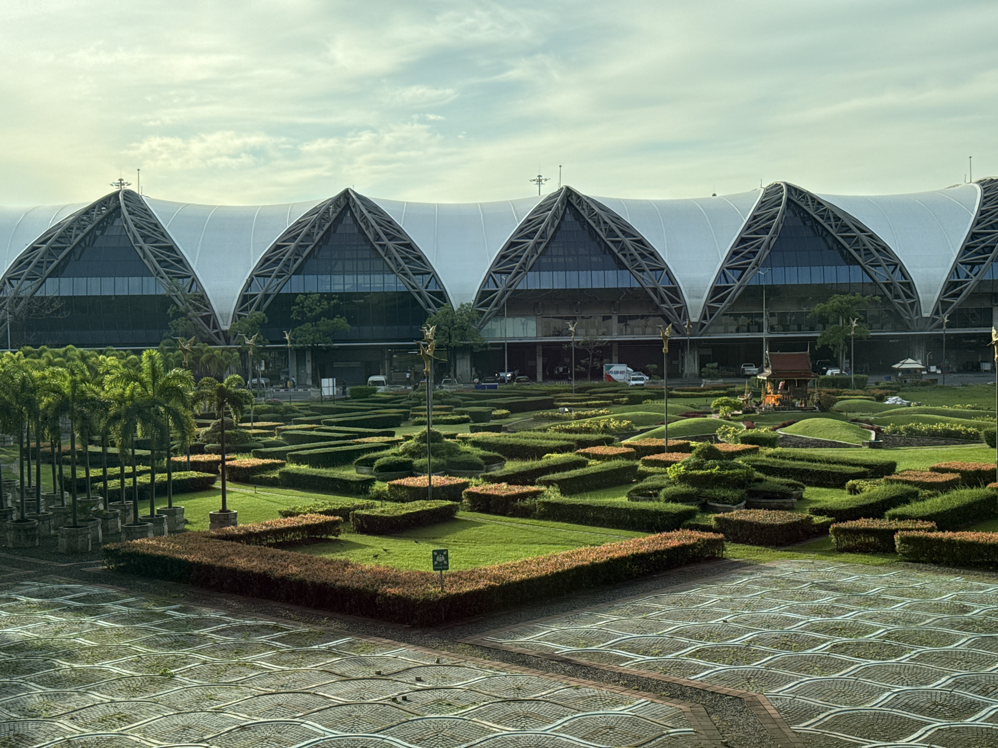 The image depicts the exterior of Suvarnabhumi Airport in Nong Prue Subdistrict, Thailand. The airport's distinctive architecture features a large, curved roof supported by a steel lattice structure, with triangular glass sections along the facade. In front of the building, an extensive, manicured garden is laid out with various shapes of hedges and rounded mounds of grass, interspersed with rows of palm trees in planters. A decorative tiled pathway with a wave-like pattern is visible in the foreground. The sky above is partly cloudy with a soft, natural light, suggesting it might be early morning or late afternoon. A small, traditional Thai-style pavilion can be seen within the garden. Several vehicles, including cars and vans, are parked or driving near the building, and a few individuals are visible in the distance. The overall scene conveys a sense of order and architectural grandeur amidst a carefully designed landscape.
