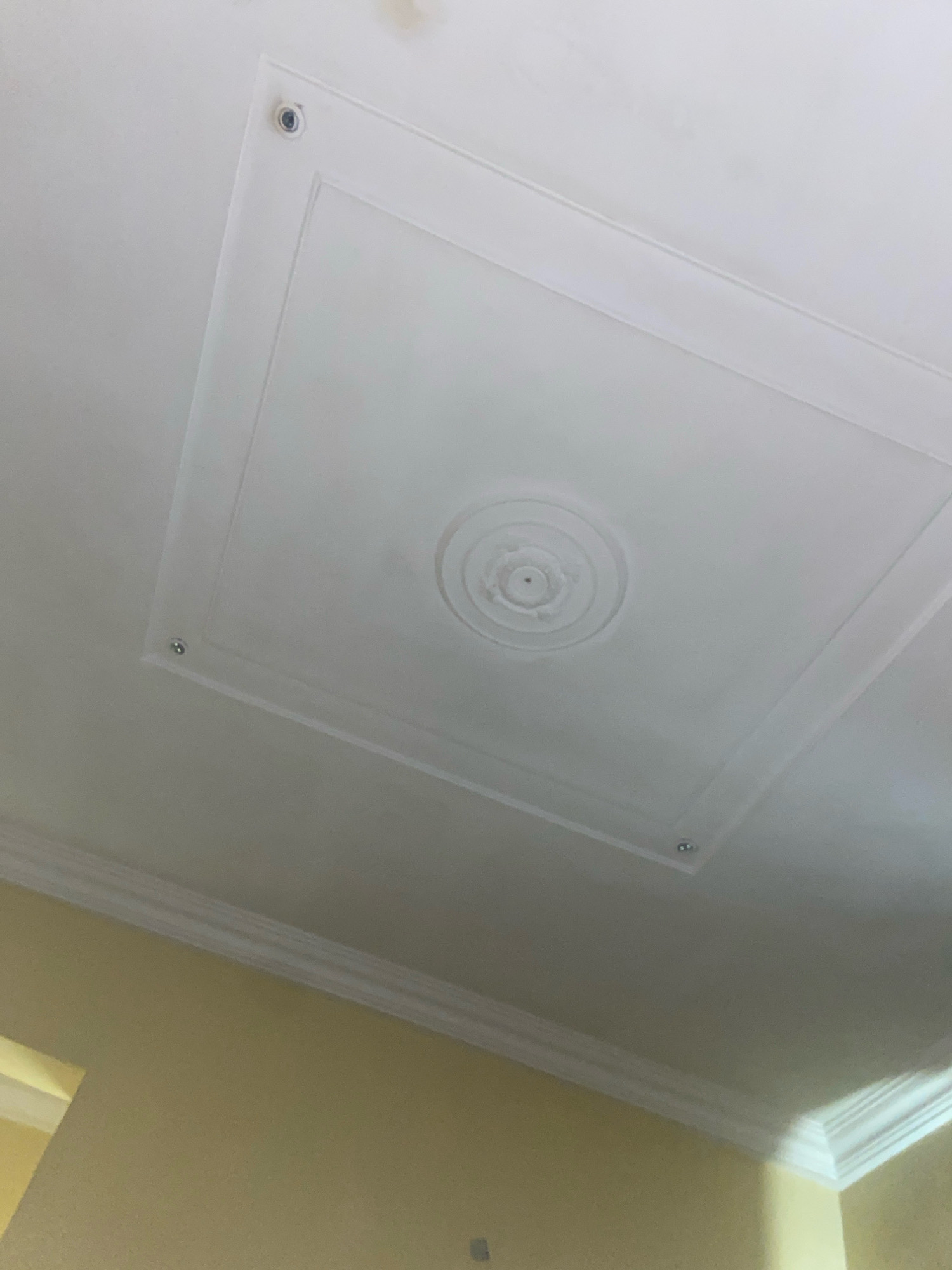 The image shows a section of a white ceiling with decorative molding. In the center of the ceiling, there is a square recessed panel with a circular, tiered design in the middle. Four small, circular fixtures or embellishments are visible near the corners of the square panel. The ceiling is lit from above, casting soft shadows. The lower portion of the image shows a light yellow wall with white crown molding where it meets the ceiling. There are no people or explicit actions depicted. The scene appears to be an interior space, possibly a room in a residential or commercial building. The location is specified as Bar Gada, Nigeria.