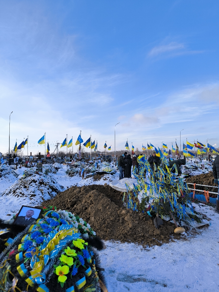 This image depicts a snow-covered section of a cemetery in Vinnytsia, Ukraine, under a clear blue sky. In the foreground, a large, dark funeral wreath adorned with numerous blue and yellow ribbons and several neon yellow artificial flowers rests on the snow. To the left of the wreath, a framed photograph of a man in military uniform is placed on the ground.
In the midground, a freshly excavated grave is visible, marked by a substantial mound of dark brown soil. Approximately five to seven individuals, dressed in dark winter attire, are gathered closely around this grave. To the right of the soil mound, a small evergreen plant or branch is heavily decorated with multiple blue and yellow ribbons.
The background reveals a large expanse of graves extending into the distance. Each visible grave is marked by a cross or headstone and features one or more Ukrainian flags (blue and yellow) mounted on poles, creating a dense field of national colors. Several streetlights are interspersed throughout the grave sites. The sky above is bright blue with a few wispy white clouds.
