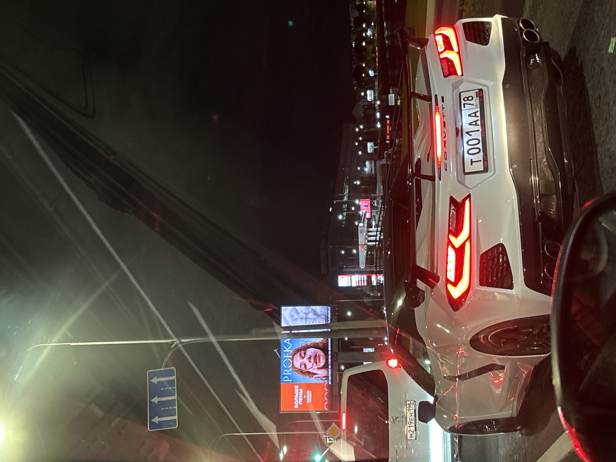 The image shows a white Chevrolet Corvette C8 with a large rear spoiler, stopped at night on a city street. The license plate reads "T001AA 78 RUS". The Corvette is in the right lane, next to a white SUV with a license plate "M219EH 198". A billboard advertising "PROFKA" is visible in the background, showing a woman with a facial mask. Traffic lights are visible, with a green light illuminated. The road appears to be wet, suggesting recent rain. The overall scene depicts a traffic stop in Saint Petersburg, Russia.