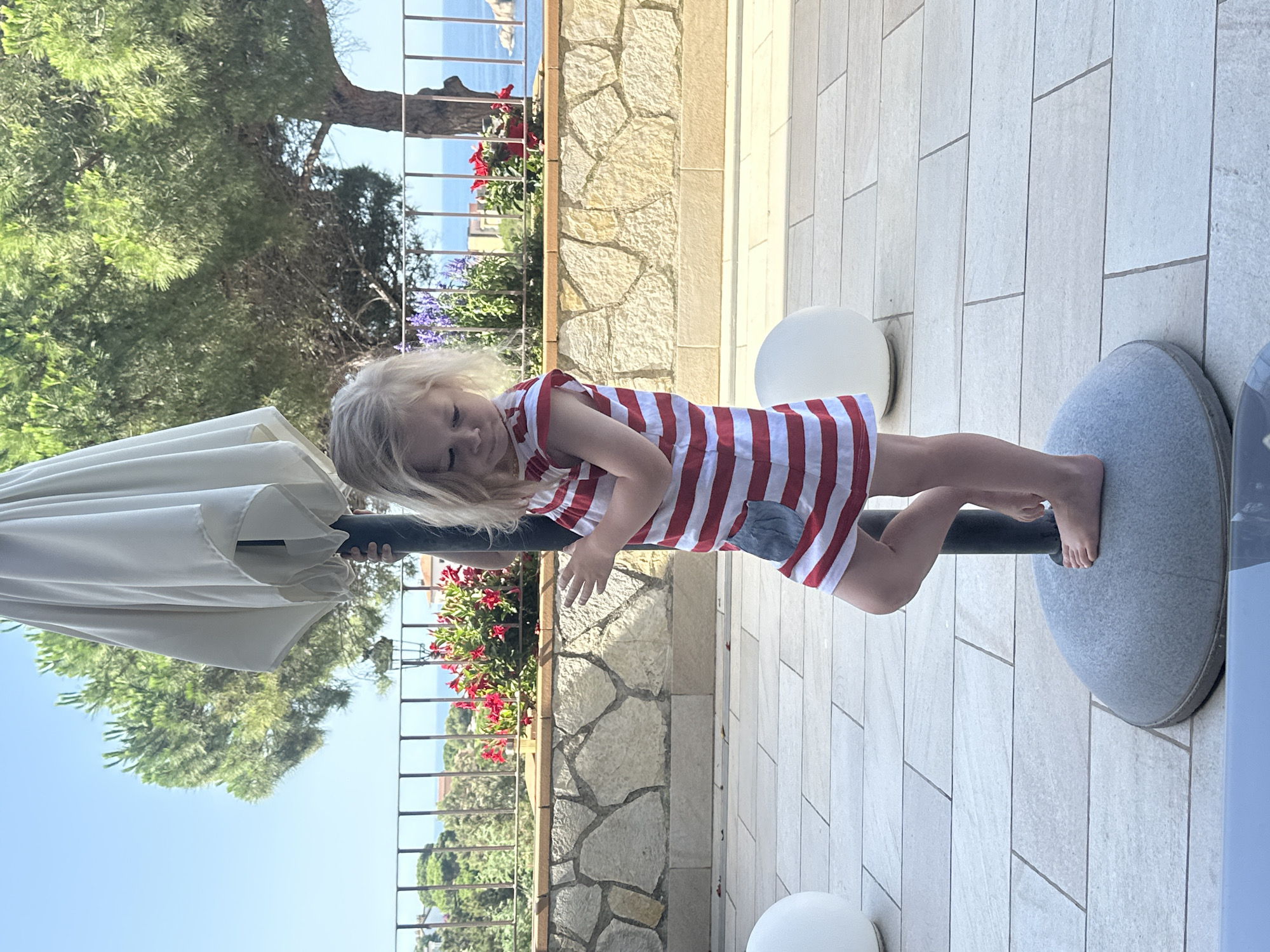 A young girl with blonde hair stands on the base of a patio umbrella, facing right. She is wearing a red and white striped dress with a grey pocket. Her left foot is placed on top of the umbrella base. The background shows a stone wall, a metal fence, flowering bushes, trees, and the sea under a clear blue sky. Two white spherical objects are visible on the tiled patio floor. The location is likely a terrace overlooking the Mediterranean Sea in Sant Feliu de Guíxols, Spain.