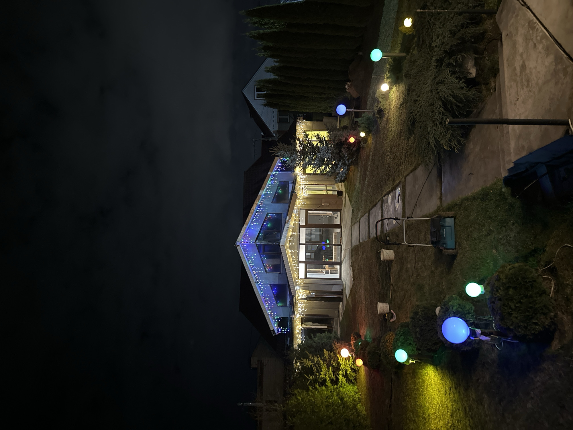 A nighttime view of a house decorated with holiday lights in an outdoor setting. The house features a prominent gabled roofline outlined with blue and white Christmas lights. String lights illuminate the front facade, and several spherical lights on posts are interspersed among the shrubbery in the yard. The lawn appears well-maintained, and a pathway leads up to the house's entrance. The sky is dark, suggesting the photo was taken at night. Based on the prompt, this scene is in Kursk, Russia.