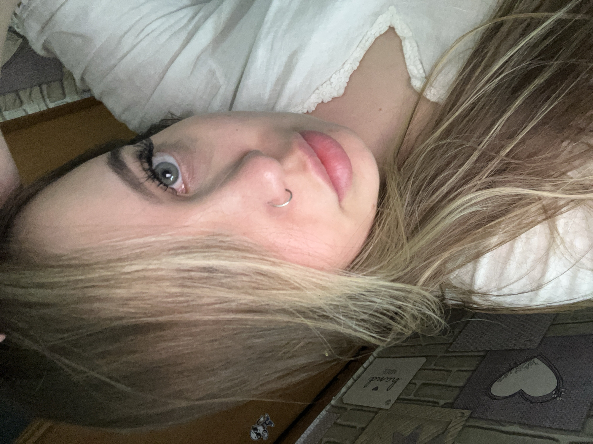 A close-up, tilted photograph depicts a young woman with fair skin, blonde and light brown highlighted hair, and light-colored eyes. She has a nose ring in her left nostril. She is wearing a white, possibly lace-trimmed garment. The background includes a gray surface with brick and heart-shaped designs, as well as a tag reading "hand made." No information suggests a location in Port-Katon, Russia. The setting appears to be indoors. The woman is lying down or reclining, with her head tilted towards the camera.