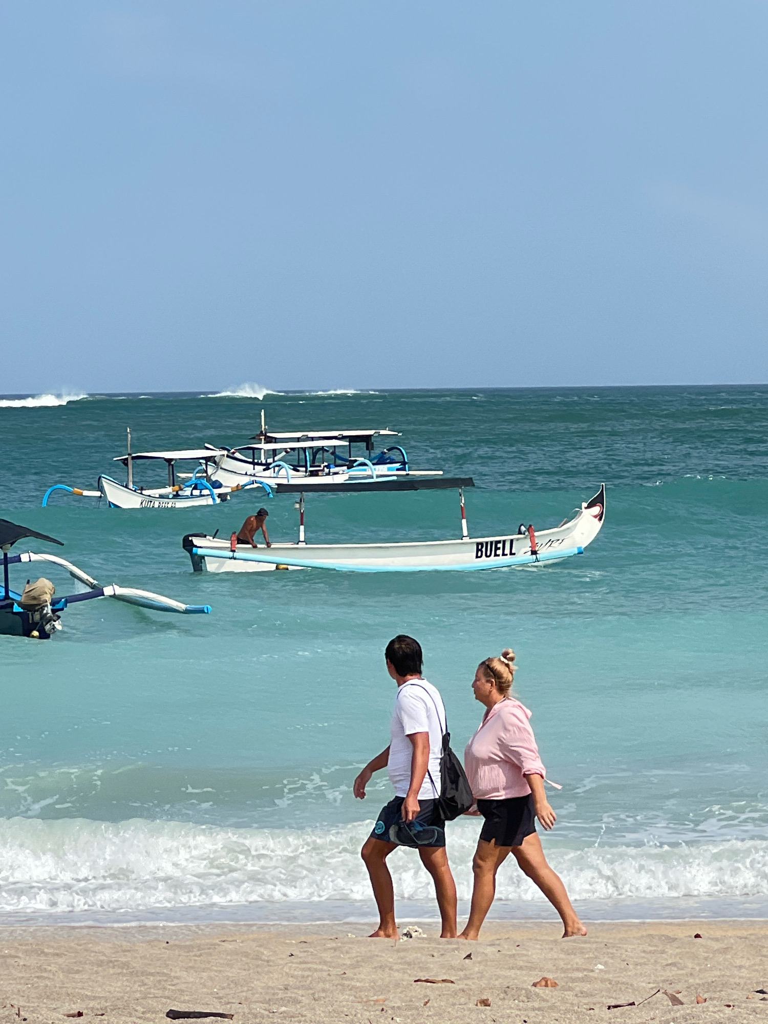 In Kuta, Indonesia, two people, a man and a woman, walk along a sandy beach. The man, wearing a white t-shirt and dark shorts, carries a pair of flip-flops. The woman, dressed in a pink hooded jacket and black shorts, has her hair in a bun and carries a black bag. In the turquoise water, several traditional Balinese fishing boats, known as jukungs, are visible. One boat prominently displays the name "BUELL." A lone figure is seen tending to a boat in the middle distance. The ocean has moderate waves breaking on the shore under a clear, light blue sky.