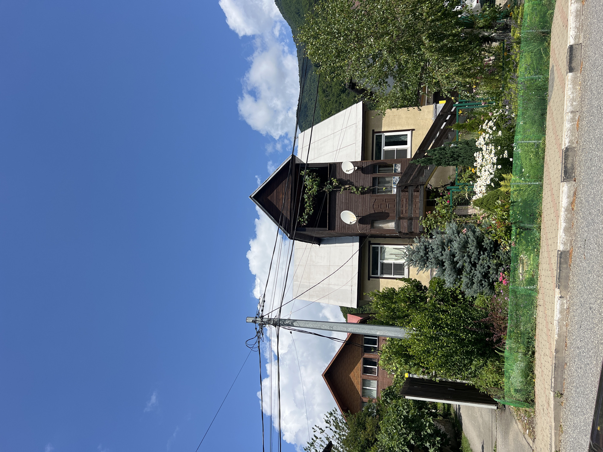 A view from a street in Krasnaya Polyana, Russia, shows a residential building with a dark brown wooden upper story and a light yellow lower story. The building has a complex roofline with several gables. A wooden staircase leads up to the upper story. There are two satellite dishes mounted on the wooden section. A small garden with various plants, including white flowers and evergreen trees, is located in front of the building, enclosed by a green mesh fence. A paved sidewalk runs along the front of the property, with a grey and red brick border. In the background, another residential building with a reddish-brown wooden facade is visible, partially obscured by trees. Overhead, utility wires are strung across a tall metal pole. The sky is blue with scattered white clouds. The image captures a sunny day in a semi-rural or suburban setting.