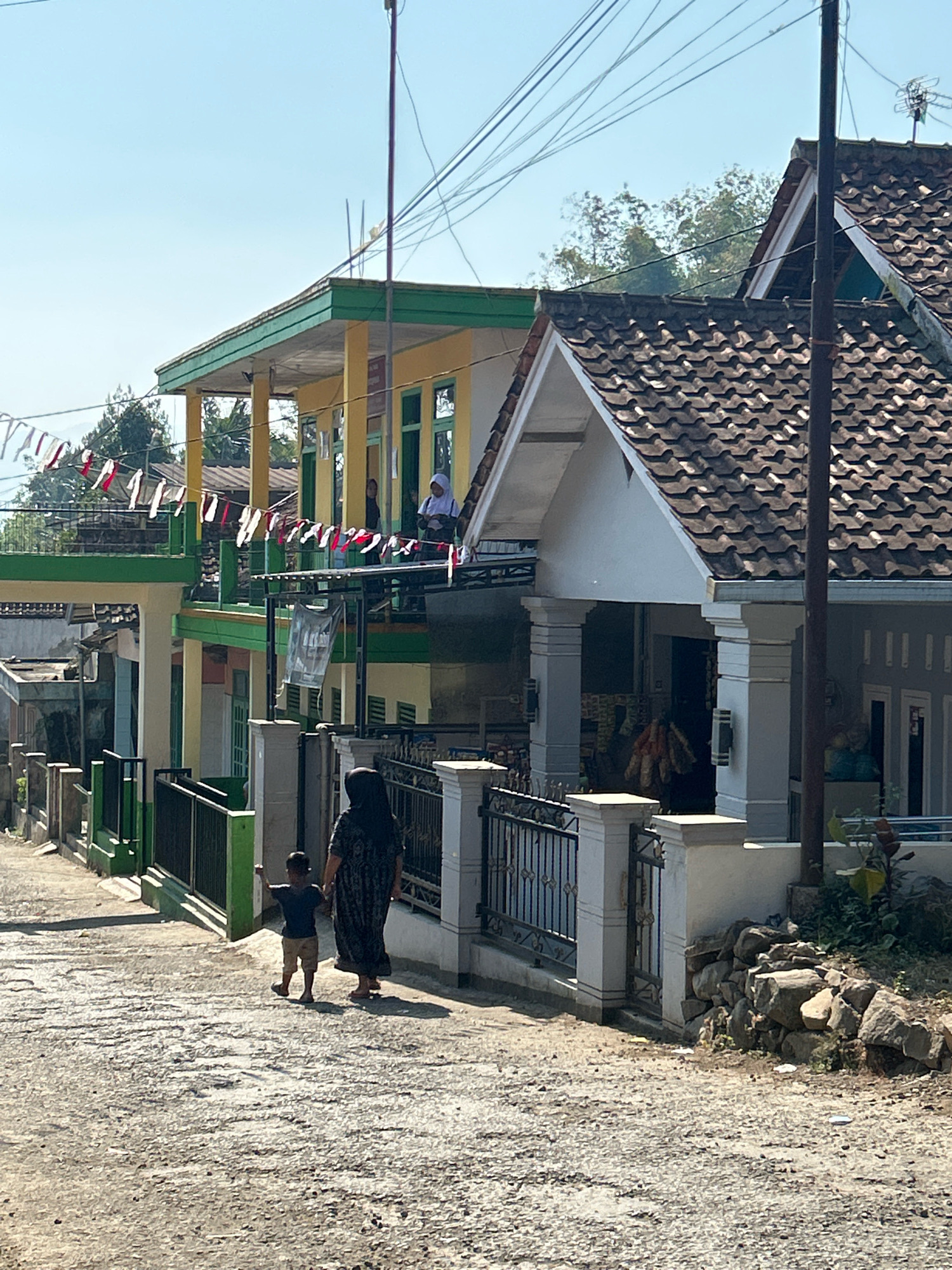 In Cihaurbeuti, Indonesia, a mother and child walk down a sunlit, uneven paved road. The mother, wearing a dark headscarf and patterned dress, holds the hand of a young boy in a dark t-shirt and tan shorts. The boy walks slightly ahead, with his left arm raised. They are walking away from the viewer, towards buildings with red and white flags strung across their balconies.
The buildings are a mix of styles. The one on the left has a yellow facade with green trim and appears to be a school or community building, indicated by the flags and people visible on the upper level. The building on the right has a grey exterior with a tiled roof and a covered entrance supported by columns. A metal fence with decorative elements runs along the property line of the grey building. In the foreground, a pile of rocks sits beside the fence. Power lines are visible crisscrossing the sky above the buildings. The scene is bathed in bright sunlight, casting shadows on the road.