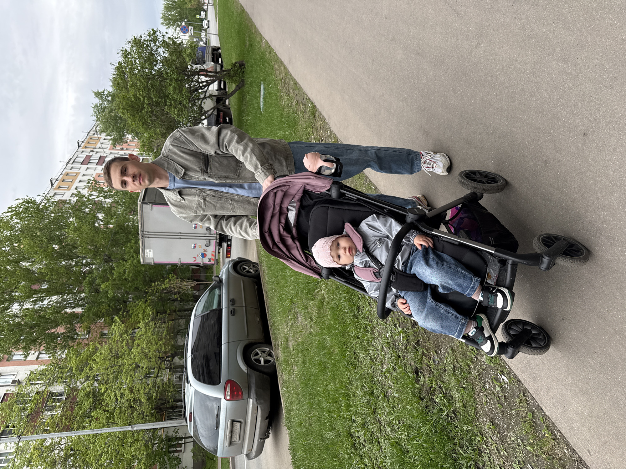 A man pushes a baby in a stroller along a sidewalk adjacent to a grassy area. The baby wears a pink hat, grey jacket, and blue jeans. The man is wearing a denim jacket and blue jeans, and white sneakers. A gray minivan and a white truck are parked nearby. The location is Moscow, Russia, as indicated by the license plate of the minivan. The ground is paved and there is a building in the background.