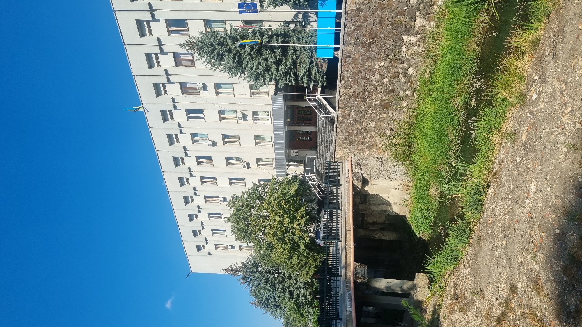 The image depicts the facade of a multi-story building with a light-colored exterior, featuring rows of windows and visible air conditioning units. A flag, appearing to be the Ukrainian flag, is visible at the top of the building. In front of the building, a wide set of stairs leads up to the entrance, flanked by railings. Several trees, including evergreen and deciduous varieties, are situated around the building and to the left of the entrance. A bridge crosses a narrow body of water, with a stone wall lining the embankment on the right side of the water. The water is bordered by lush green grass and reeds. The scene is bathed in sunlight, casting shadows on the ground and the water. The overall impression is of an institutional building situated in a semi-natural, urban environment. This building is identified as the Berehove City Council building in Berehove, Ukraine.