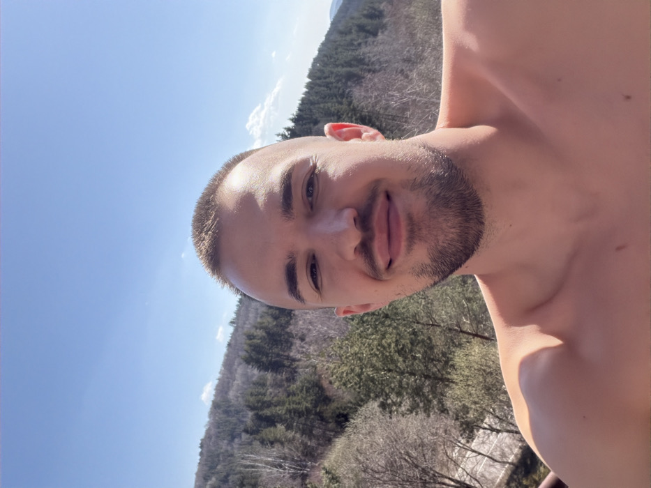 An adult male subject is depicted in a close-up outdoor selfie, bare-chested and facing the camera with a slight, closed-lip smile. He has short, light-colored hair and a short beard. The background features a mountainous, forested landscape under a clear blue sky with minimal white clouds visible towards the upper right. Dense evergreen and bare deciduous trees cover the slopes. A light-colored, winding road is visible in the lower-left background, traversing the forested area. The scene is consistent with the mountainous terrain near Kladnitsa, Bulgaria.