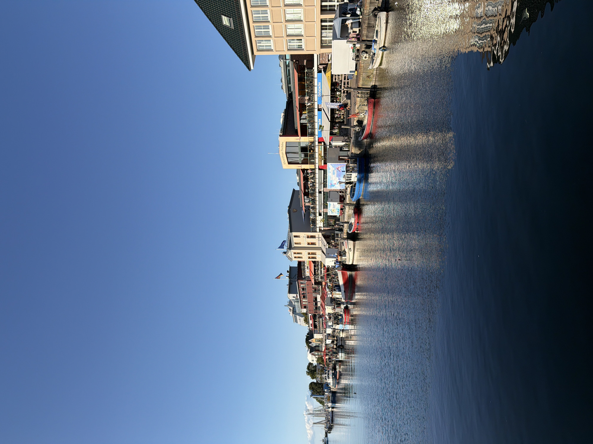 This is a horizontal, eye-level shot of the Warnow River in Rostock, Germany, specifically the harbor area. The sky is a clear, bright blue, with a few faint clouds on the far left. Along the riverbank, there is a row of buildings with shops and restaurants, many with outdoor seating covered by red awnings. Several boats are docked along the pier, including fishing boats and smaller pleasure craft. The buildings are reflected in the calm water of the river, creating a shimmering effect. The scene appears to be set in the late afternoon, as evidenced by the warm light and long shadows. There are a few people visible walking along the pier or sitting at the outdoor cafes, but the overall atmosphere is peaceful and serene. The architecture of the buildings suggests a maritime theme, with some featuring nautical flags and signage.