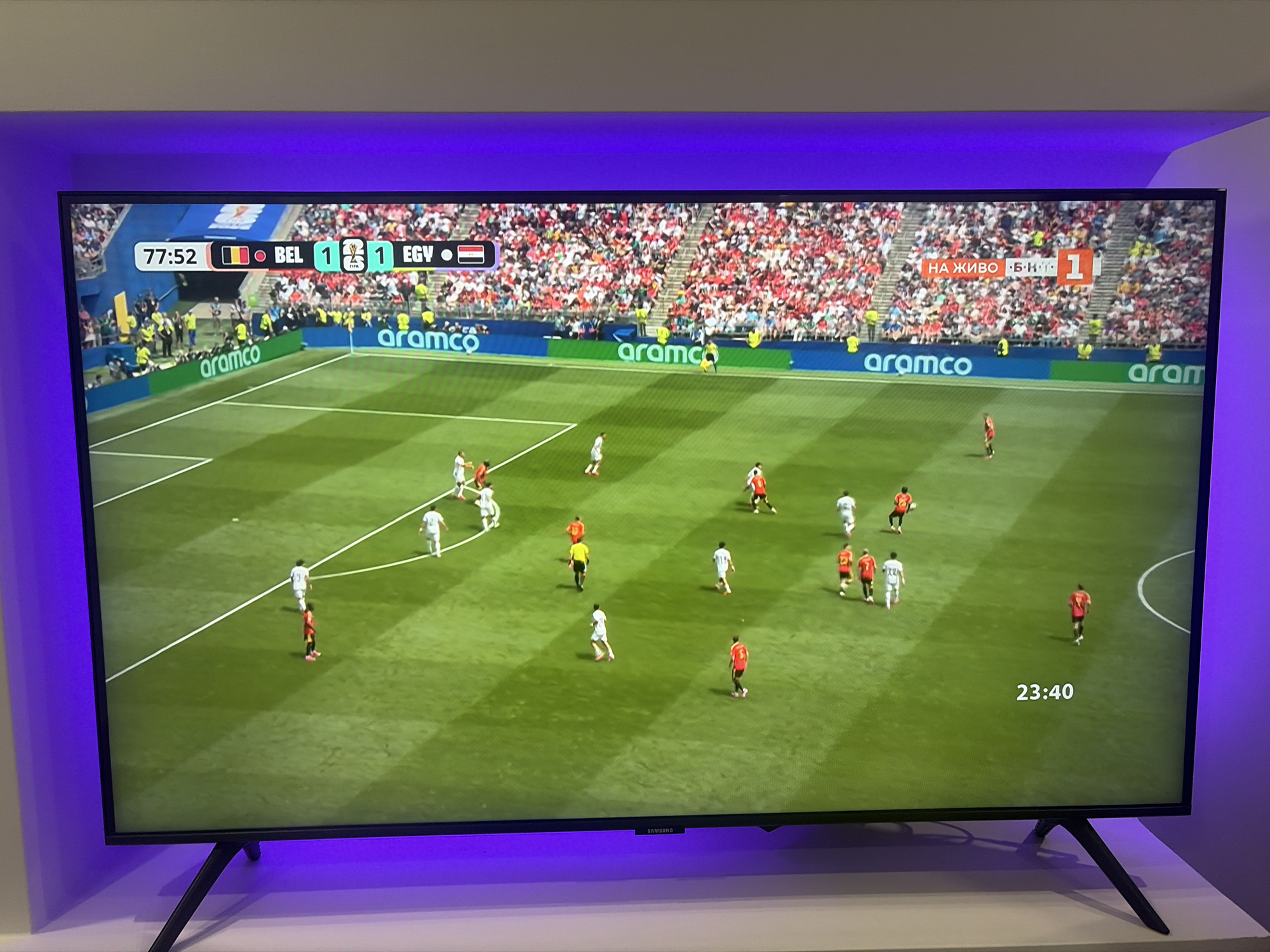 The image shows a television screen displaying a soccer match. The score is 1-1 between Belgium (BEL) and Egypt (EGY). The time is 77:52. The game is being broadcast live, as indicated by the text "НА ЖИВО" (meaning "live" in Russian) in the upper right corner, with a score of 1-1. The field is green with white markings. Several players in red and white uniforms are visible on the field, in various positions indicating ongoing play. The stadium seating is visible in the background, filled with spectators. The TV screen is framed by purple LED lighting. The television is placed on a white stand with black legs. The lower part of the image shows the bottom of the TV screen and the stand. The television has the "SAMSUNG" logo on its bottom bezel. There is no visible indication of the location being in the city of Podgumer, Bulgaria.