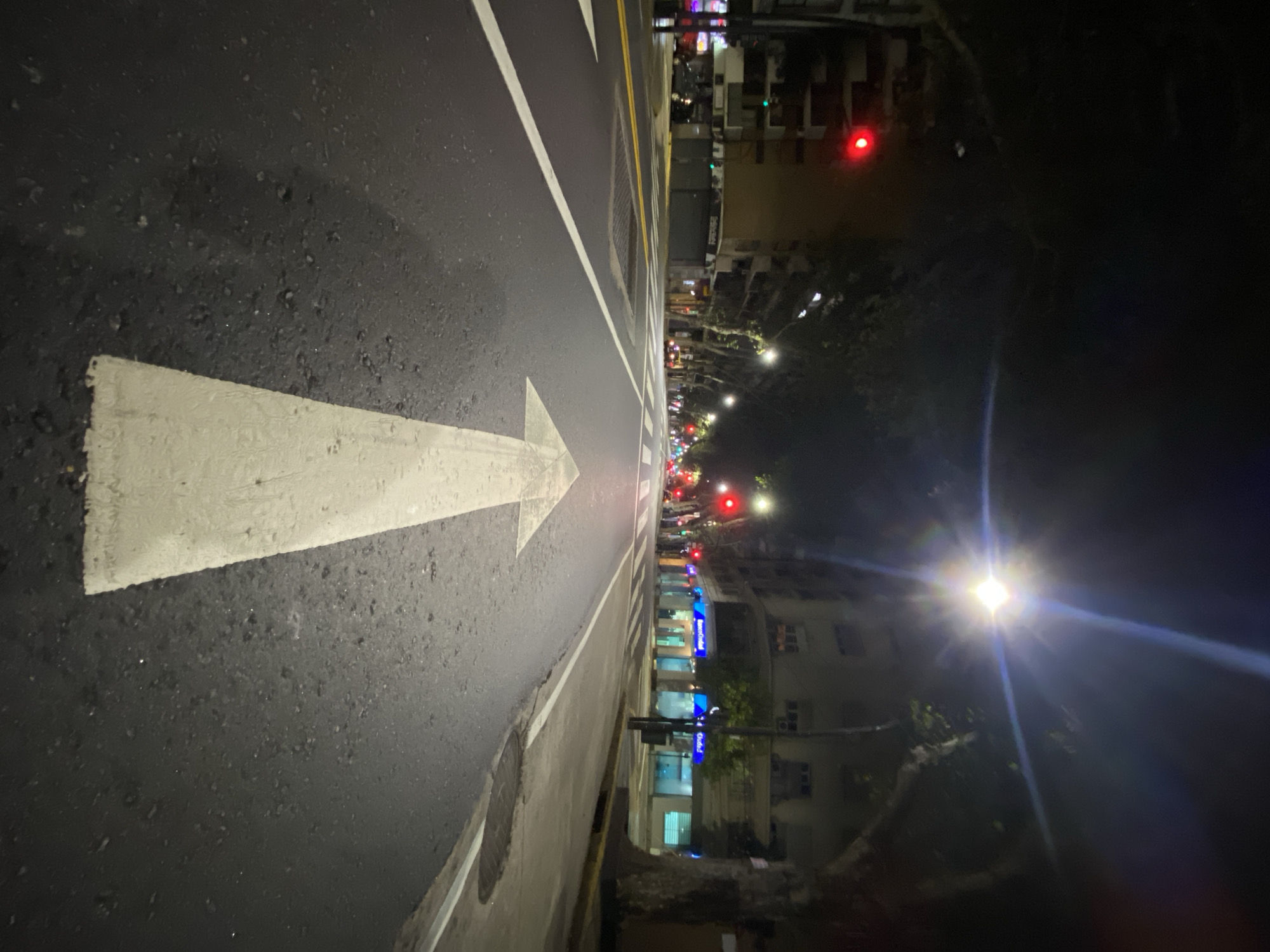 At night, a street view shows an asphalt road with a white painted arrow pointing forward. The right side of the road features buildings with illuminated windows and streetlights, with one light source producing a visible lens flare. Traffic lights are visible in the distance, showing red. Based on the location of the “Banco Ciudad” storefront sign, this scene is in Buenos Aires, Argentina.