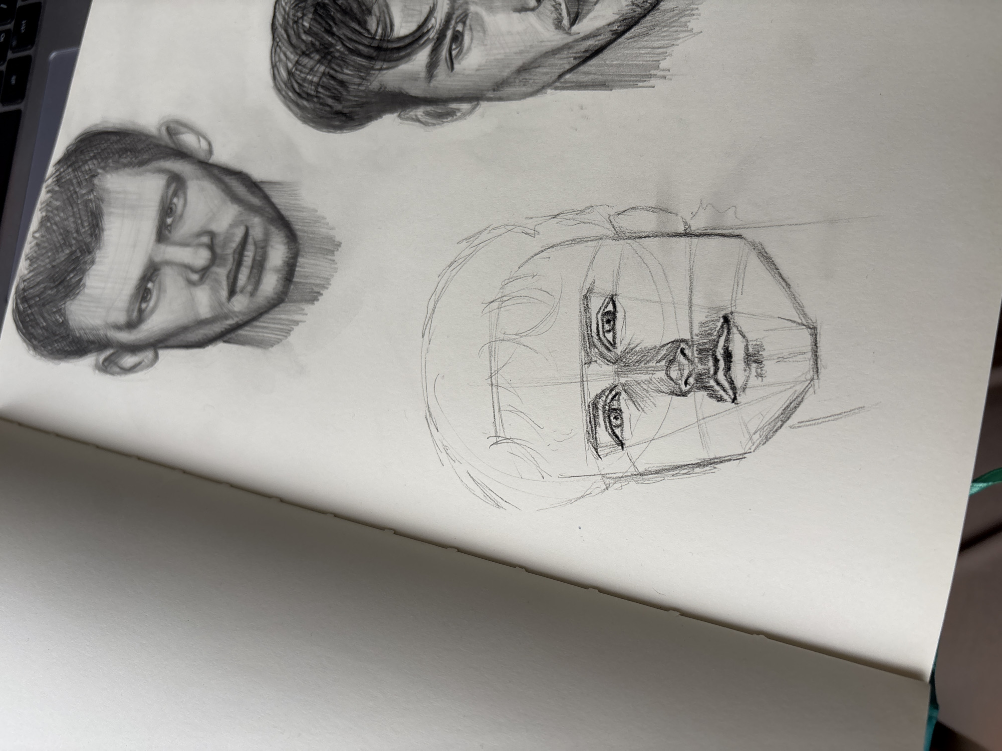 The image shows a sketchbook lying open. On the right page are three pencil sketches of faces. The top sketch is a partially shaded male face with dark hair. The middle sketch is a fully shaded male face with short hair. The bottom sketch is a partially drawn male face with guidelines sketched in. The environment is a room with a notebook and laptop. Izhevsk, Russia is not present.