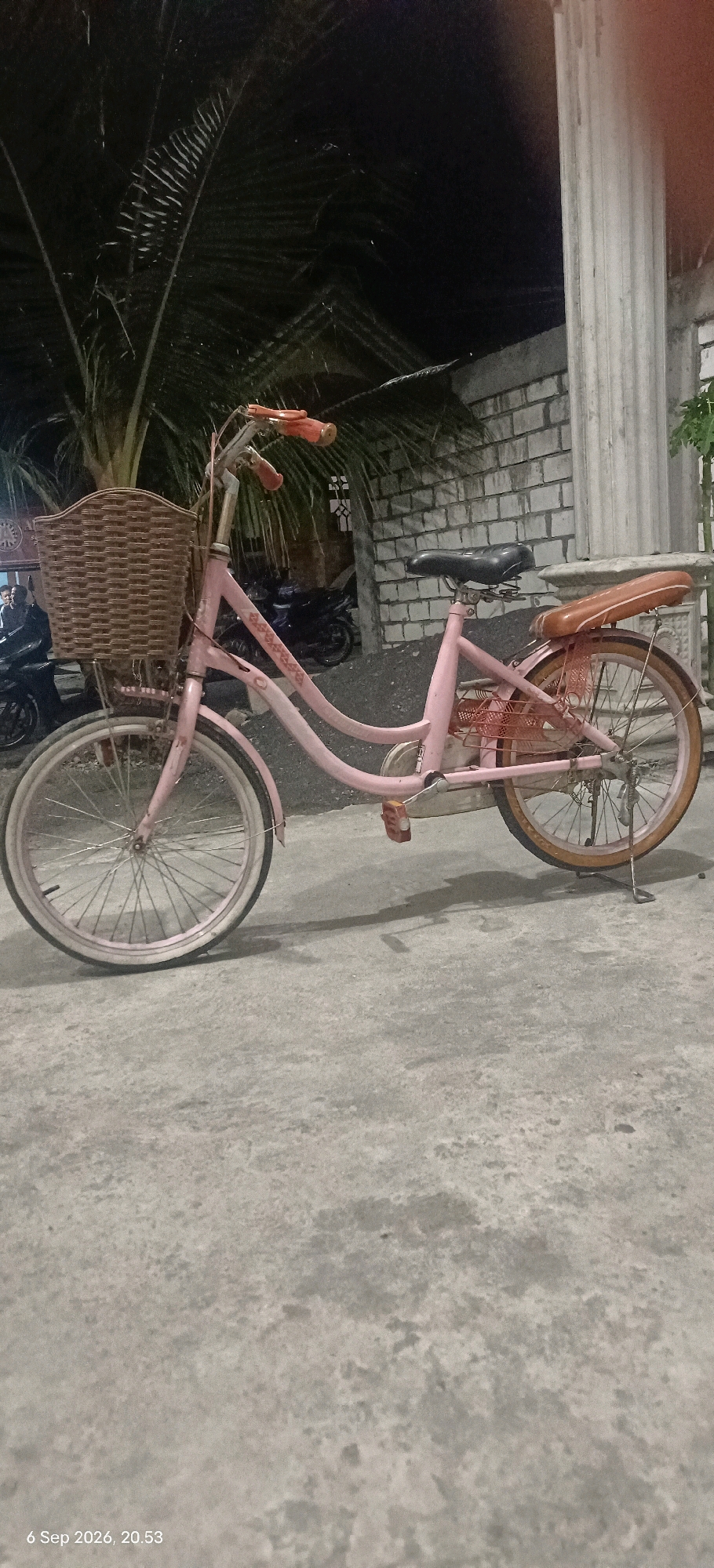 A pink bicycle with a brown woven basket is parked on a concrete surface at night. The bicycle has a brown saddle and a brown rack over the rear wheel. Behind the bicycle, a palm tree is visible, as well as a brick wall and a white column. In the background, two motorcycles are parked. The image includes a timestamp in the lower left corner: "6 Sep 2026, 20.53". The location is identified as Mojopetung, Indonesia.