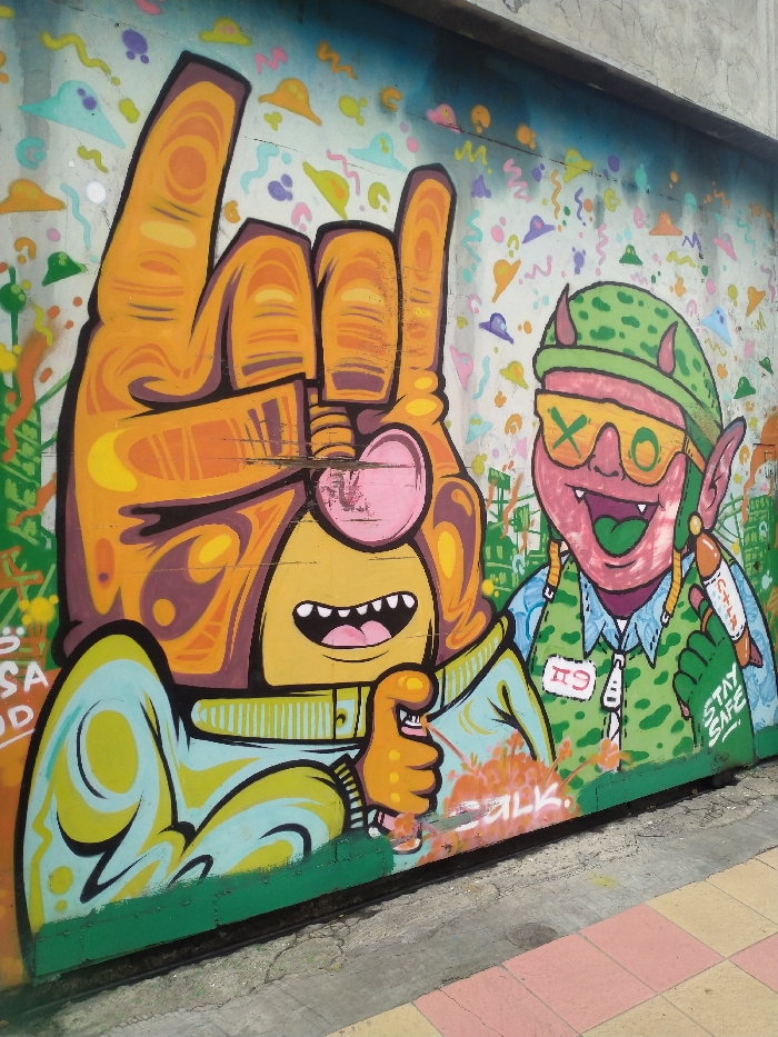 A vibrant graffiti mural adorns a concrete wall, featuring two stylized, cartoon-like characters rendered in bold outlines and bright colors. The scene is brightly lit, suggesting daytime, and the base of the wall transitions to a concrete path with pink and yellow checkered tiles, indicating an outdoor urban setting in Karanganyar, Indonesia.
On the left, a large, orange, gloved hand forms a "rock on" or "I love you" gesture with its thumb, index, and pinky fingers extended. Below this hand, a character with a yellow face, wide open-mouthed smile, and visible teeth is depicted. A pink heart shape partially covers its eye area. This character wears a light blue and yellow long-sleeved garment with swirling patterns.
To its right, a green-skinned imp or goblin-like character is portrayed. It has pointed ears and small red horns protruding from a green, camouflage-patterned military-style cap. Large yellow sunglasses obscure its eyes, bearing the text "X O" on the lenses. This character also has a wide, joyous, open-mouthed expression with a green tongue and visible teeth. It wears a camouflage vest over a light blue shirt, with a white tag pinned to its chest displaying "#9". In its right hand, it holds a red, rocket-shaped object, possibly a spray can or firecracker, with the text "CITY" and "STAY SAFE" visible on its side.
The background of the mural is a lighter wall covered with numerous small, colorful, abstract shapes, including stylized clouds, squiggles, and other organic forms, giving a playful and whimsical feel. Near the bottom center of the mural, below the right character, faint orange paint splatters partially obscure the word "SILK".