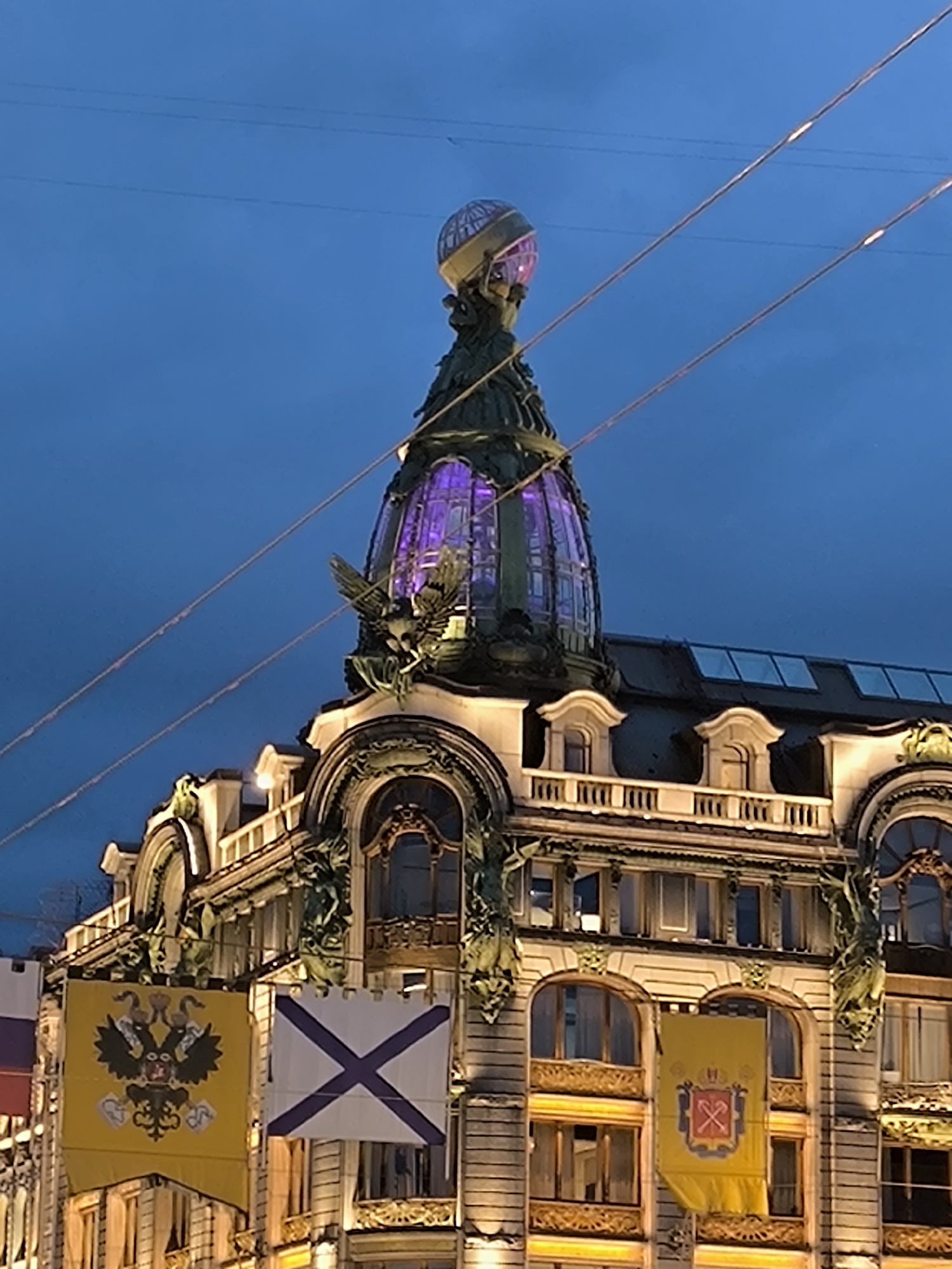 This image captures the upper portion of the Singer House (Dom Knigi), an Art Nouveau architectural landmark located on Nevsky Prospekt in Saint Petersburg, Russia, at what appears to be twilight or night.
The central feature is the building's distinctive glass dome, illuminated with a vibrant purple light. Atop the dome, a large, translucent globe is visible. The dome's base is adorned with ornate sculptural details, including two winged figures resembling eagles or mythical birds with outstretched wings. The building's facade below the dome is intricately detailed with classical and Art Nouveau elements, including arched windows, balconies with balustrades, and decorative scrollwork. Warm yellow artificial lighting illuminates the building's architectural features, contrasting with the dark blue, overcast sky.
Three large banners hang from the building's facade. The leftmost banner is yellow and displays a black double-headed eagle with crowns, an orb, and a scepter, characteristic of the historical Imperial Standard of Russia. The middle banner is white with a blue diagonal cross (Saltire of St. Andrew), representing the Russian Naval Ensign. The partially visible banner on the right is yellow and features a crowned red shield containing crossed silver hammers and picks, a historical emblem often associated with mining or industry.
Two thin, light-colored parallel lines, likely overhead wires or cables, traverse diagonally across the upper left and central portions of the dark blue sky. No individuals are visible in the frame.