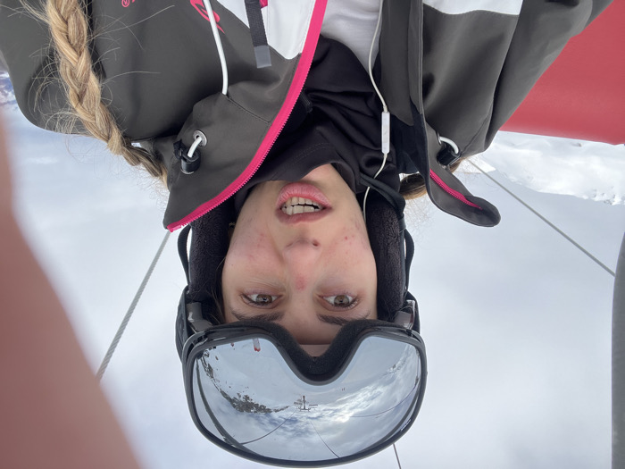 A selfie-style portrait captured on a ski lift shows a young woman, rotated for proper viewing. She is depicted from the upper chest upwards, with light brown or blonde hair styled in a braid that falls over her left shoulder. Her eyes are open, looking directly at the camera, and her mouth is slightly agape, revealing her teeth, with a neutral to light expression. She has some visible blemishes on her skin.
She is dressed in winter sportswear, including a dark olive green or grey ski jacket with white sections and bright pink trim on the zippers and edges. Beneath this, a black hooded layer and a white base layer are visible. White wired earphones are in her ears, with the wire draped over her jacket. A black ski helmet with fleece ear pads covers her head, and dark-framed ski goggles with reflective lenses are pulled up onto her forehead, obscuring her eyebrows. The goggles distinctly reflect a snowy mountain landscape and parts of a ski lift system, including cables or pylons.
The background shows an overcast sky and snow-covered terrain, characteristic of a high-altitude winter environment. To her right, a bright red component, likely part of the ski lift chair, is visible. A thin white rope or cable extends upwards on her left side, possibly part of the lift's structure or a safety element. The scene suggests a casual moment during a skiing trip in Val-d'Isère, France. No discernible text is visible on her clothing or equipment.