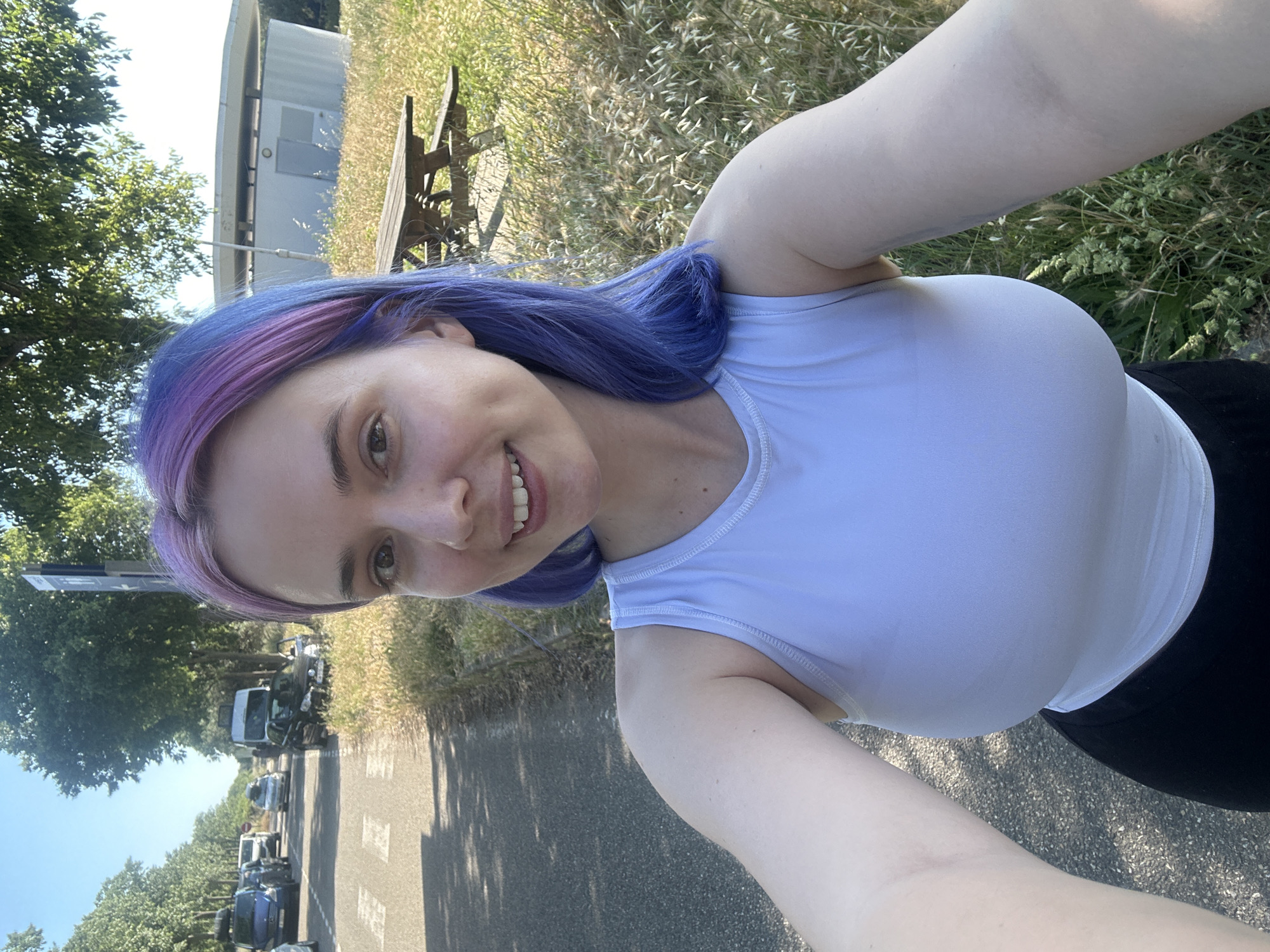 A young woman with purple and blue dyed hair smiles into the camera. She is wearing a light blue tank top and black bottoms. She appears to be on a road or path, with parked cars visible in the background, along with trees and green foliage. The location is Caissargues, France. A building and a wooden bench are also visible in the background.