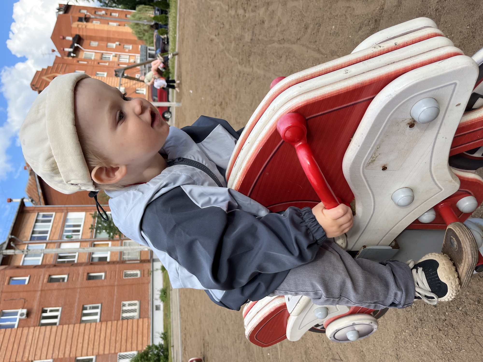 A young child, likely a toddler, is captured playing on a red and white spring rider in an outdoor playground located in Chaikovsky, Russia. The child is seated on the playground equipment, holding onto its red handles, and appears engaged in the activity, looking upwards with a slightly open mouth, suggesting curiosity or enjoyment.
The child is dressed in a light-colored cap, a two-toned grey and dark blue jacket, grey pants, and dark blue and light-colored sneakers. The spring rider features a dominant red surface with white accents and grey elements, showing some signs of wear.
The background reveals a sandy ground typical of playgrounds. Beyond the play area, several multi-story residential buildings constructed of brick-colored material are visible. These buildings feature numerous windows and hint at an urban or suburban setting. The sky is bright blue with scattered white clouds, indicating a clear and sunny day, likely during late morning or afternoon. The overall atmosphere conveys a moment of childhood play and leisure in a residential environment. No visible text is present in the image.