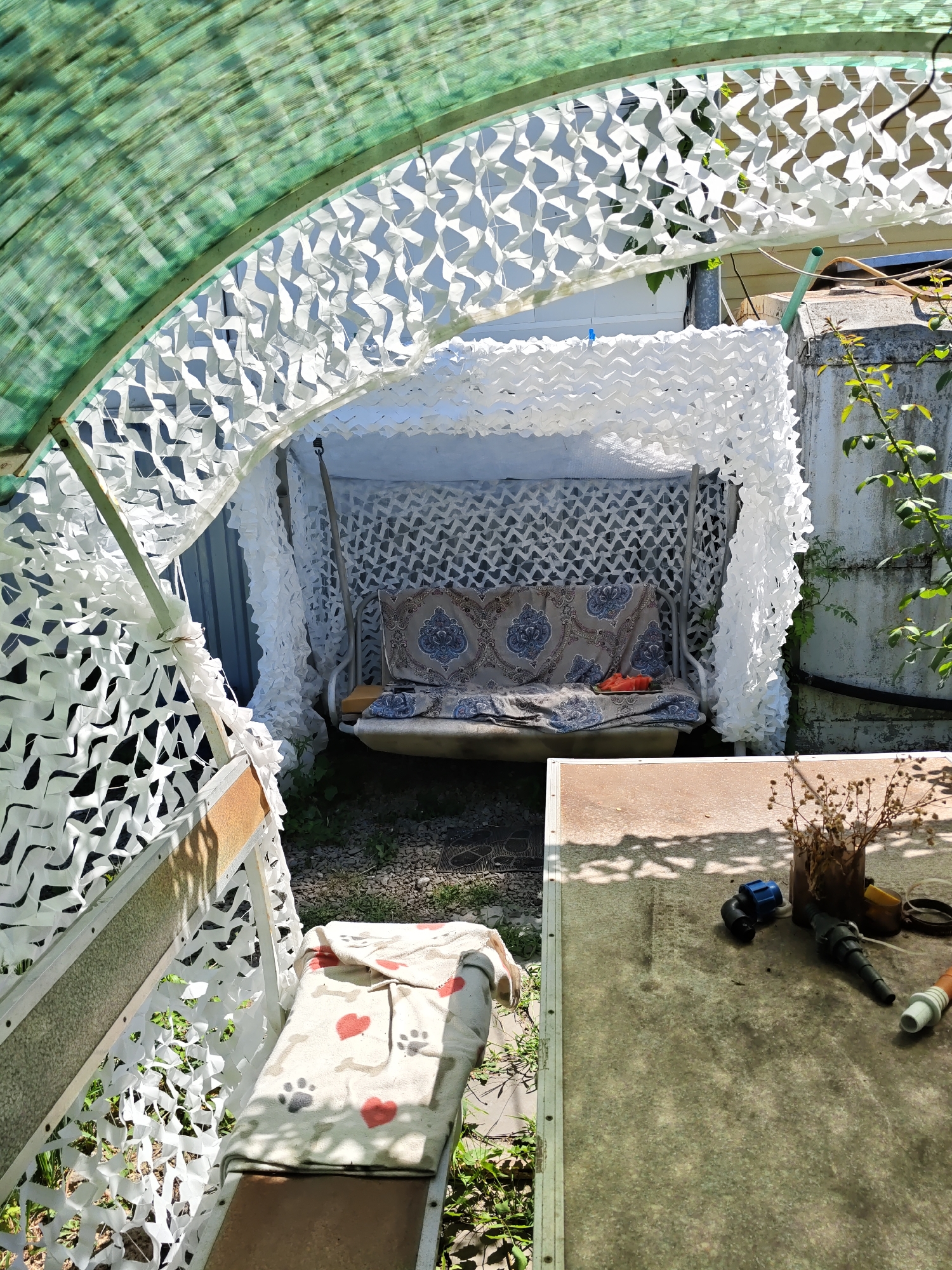 The image depicts a partially covered outdoor seating area, likely a garden or backyard, with a swing set and a table. The area is shaded by a green, translucent canopy, and the swing set is further concealed by white camouflage netting. The swing itself has a cushion with a patterned fabric. To the left, a bench is covered with a blanket adorned with hearts and paw prints. On the table in the foreground, various objects are scattered, including what appears to be gardening or plumbing tools, and a small vase with dried flowers. The overall impression is of a functional, yet somewhat cluttered, outdoor space. The location is identified as Rostov-on-Don, Russia.