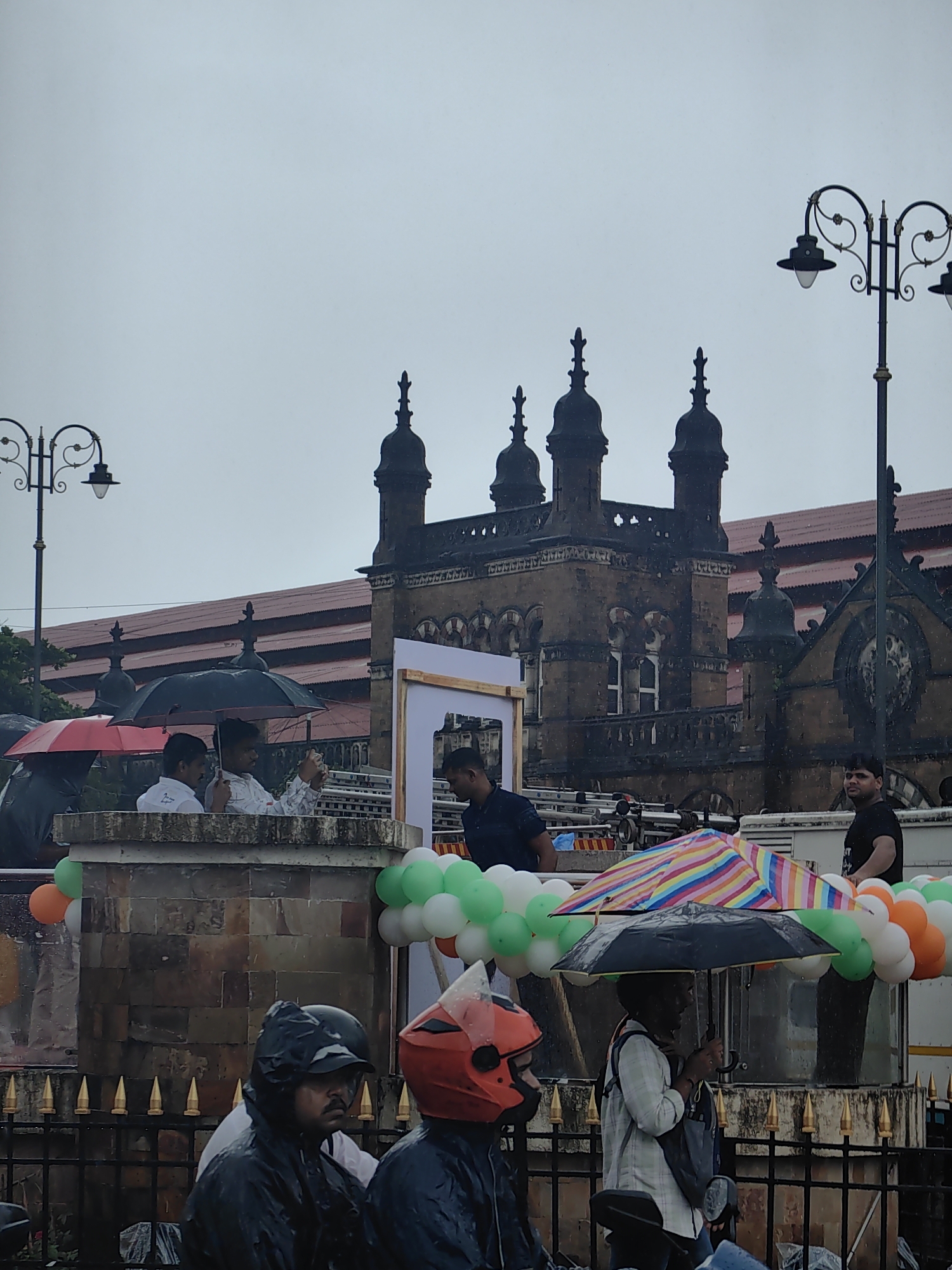The image shows a partially rainy day scene in front of the Chhatrapati Shivaji Maharaj Terminus (CSMT) in Mumbai, India. Several individuals are present, some under umbrellas, suggesting precipitation. In the foreground, people are on motorbikes, one wearing a red helmet and another a black helmet and a dark rain jacket. Behind them, a person in a light-colored shirt is holding a colorful, striped umbrella over themselves and another individual who is out of frame. Further back, a group of people are standing behind a low wall decorated with balloons in the colors of the Indian flag (green, white, and orange). The iconic architecture of CSMT, with its intricate stone carvings and domed structures, is visible in the background under a cloudy sky. A street lamp with multiple lights is also visible on the left side of the image.