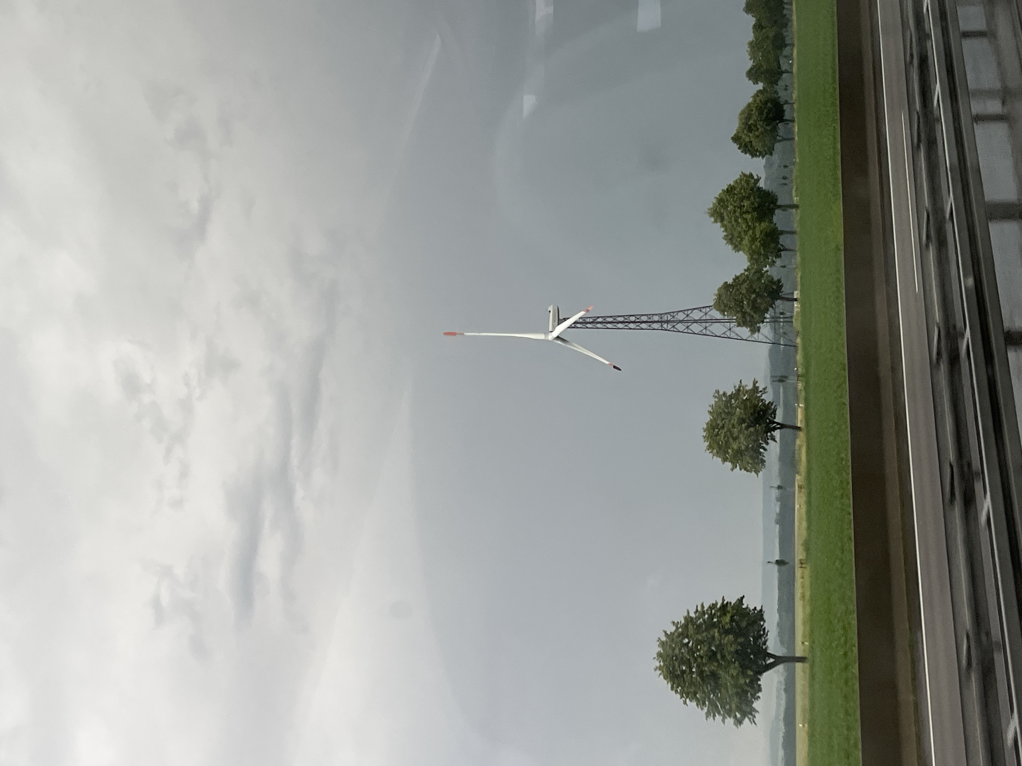 The image shows a landscape scene with a wind turbine in the background, located near Pohle, Germany. The sky is overcast and appears to be a stormy grey. In the foreground, there is a highway with a wet surface, reflecting the sky. A green grass field runs alongside the highway, with a row of trees forming a line in the distance. The wind turbine is a prominent structure, with its blades positioned upwards. The overall atmosphere is somber and perhaps indicative of impending rain.