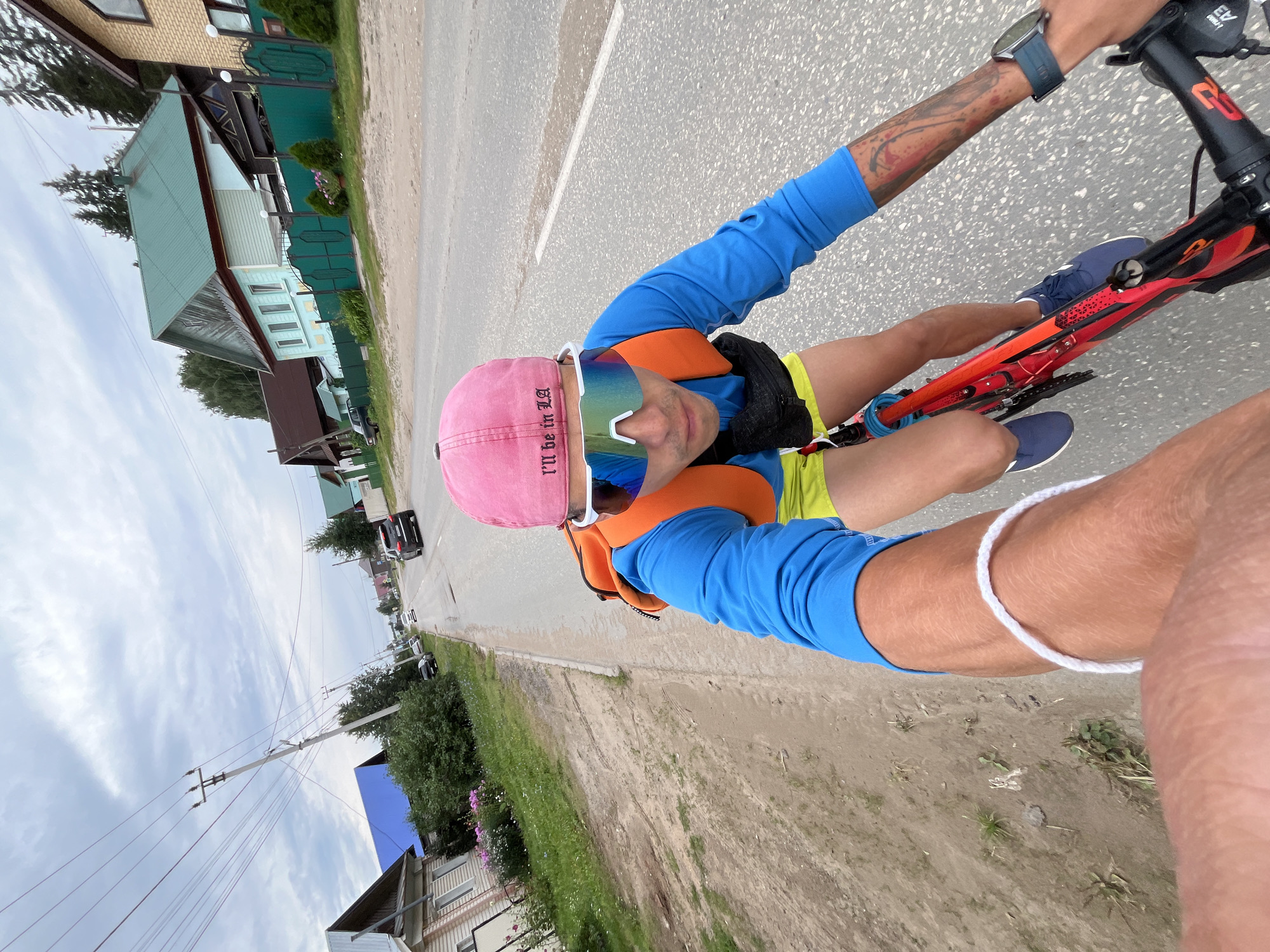 A person is riding a red bicycle on a paved road in a rural or suburban area. The person is wearing a blue long-sleeved shirt, yellow shorts, a pink baseball cap with the words "I'll be in LA" embroidered on it, and mirrored cycling sunglasses. They have an orange backpack on and a black fanny pack around their waist. The road is flanked by a dirt shoulder and green grass. Several houses with gabled roofs and a green fence are visible on the right side of the road, characteristic of residential areas in Chaikovsky, Russia. Power lines run overhead. The sky is overcast.