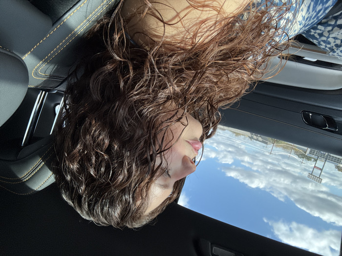 The image depicts a woman with wet, curly, brown hair, seen from a low angle inside a car. Her face is tilted upward, showing her lips and nose. The car's interior features a black dashboard with yellow stitching. Outside the car window, a clear blue sky with fluffy white clouds is visible. A building or structure with tall poles appears in the distance, reflected in the window.