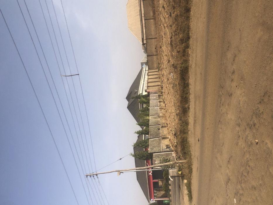 A street view showcases a dirt road alongside a building with a dark roof and a row of solar panels. Another building is visible in the background. Above, a power pole supports a series of electrical wires. A stick is precariously perched on one of the wires. The sky is pale blue.