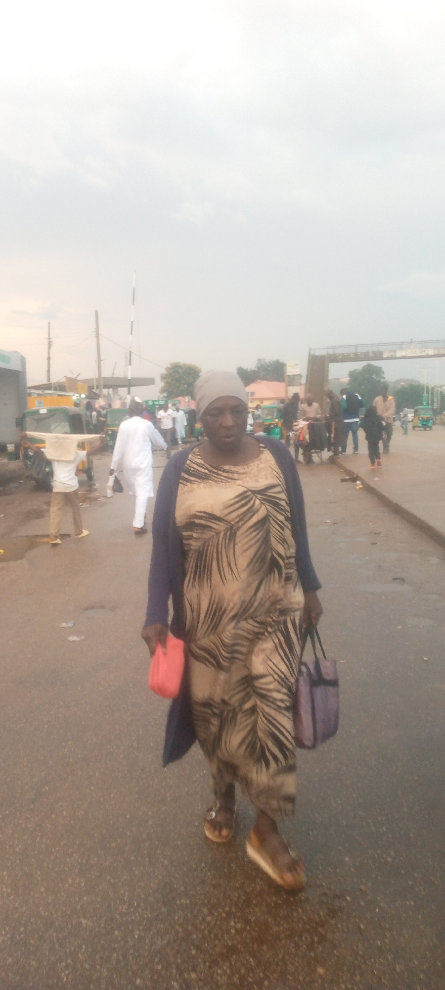 A woman in a patterned dress and blue cardigan walks across a paved road in an outdoor market setting. She is wearing a light gray headscarf and appears to be holding a red object in her left hand and a purple bag in her right. The road is wet, suggesting recent rain. In the background, there are several auto-rickshaws and other people walking. The sky is overcast.