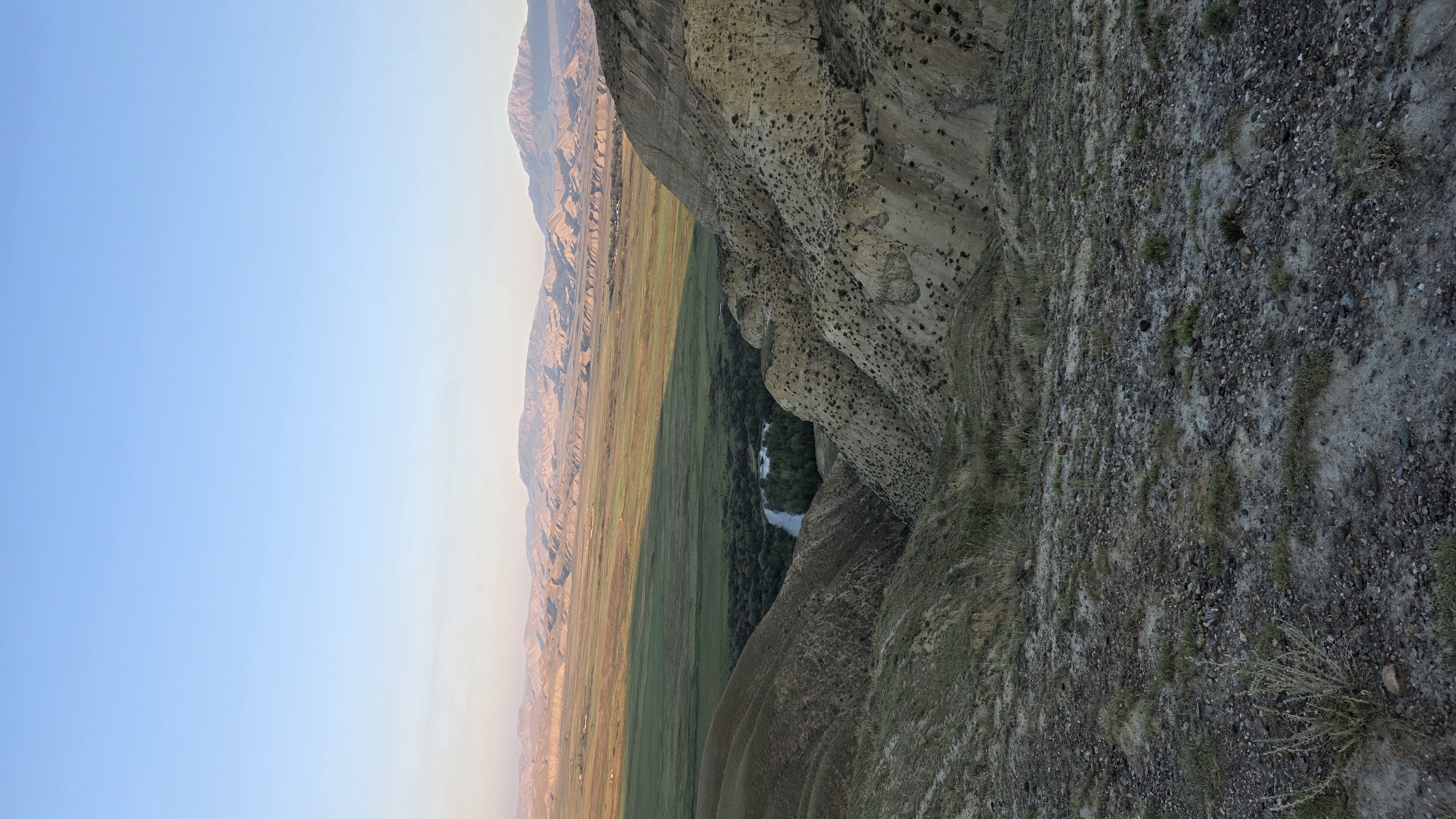 A wide shot captures a natural landscape, likely in or near Ак Жар, Kyrgyzstan, under a clear, pale blue sky. The foreground is dominated by a rocky, sparsely vegetated hillside, with dry grasses and small stones scattered across a sandy, light-colored soil. This slope descends into a valley.
Further in the middle ground, a verdant green field stretches out, bisected by a winding river lined with a dense growth of trees. The river reflects the sky and is a prominent feature in the valley. The sunlight casts a distinct shadow across the green field, indicating the direction of the sun.
In the background, a series of rolling hills and distant mountains form the horizon. These landforms exhibit layered geological strata and appear arid and weathered, bathed in the soft light of either sunrise or sunset, which lends a warm, diffused glow to their surfaces. The overall scene evokes a sense of vastness and natural tranquility, with no human presence visible.