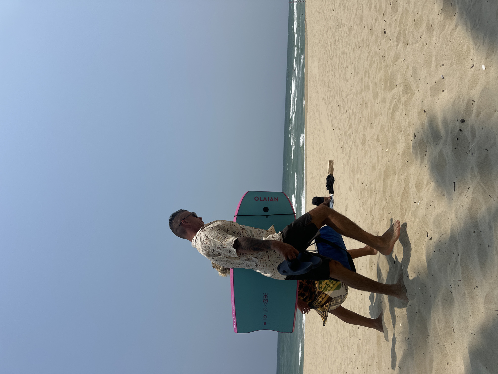 On a sandy beach in Đà Nẵng, Vietnam, two people stand near the ocean. One person is carrying two turquoise and pink boogie boards labeled "OLAIAN." The individuals are dressed in beach attire with one in dark shorts and a light patterned shirt, and the other partially visible in patterned shorts and a blue shirt.