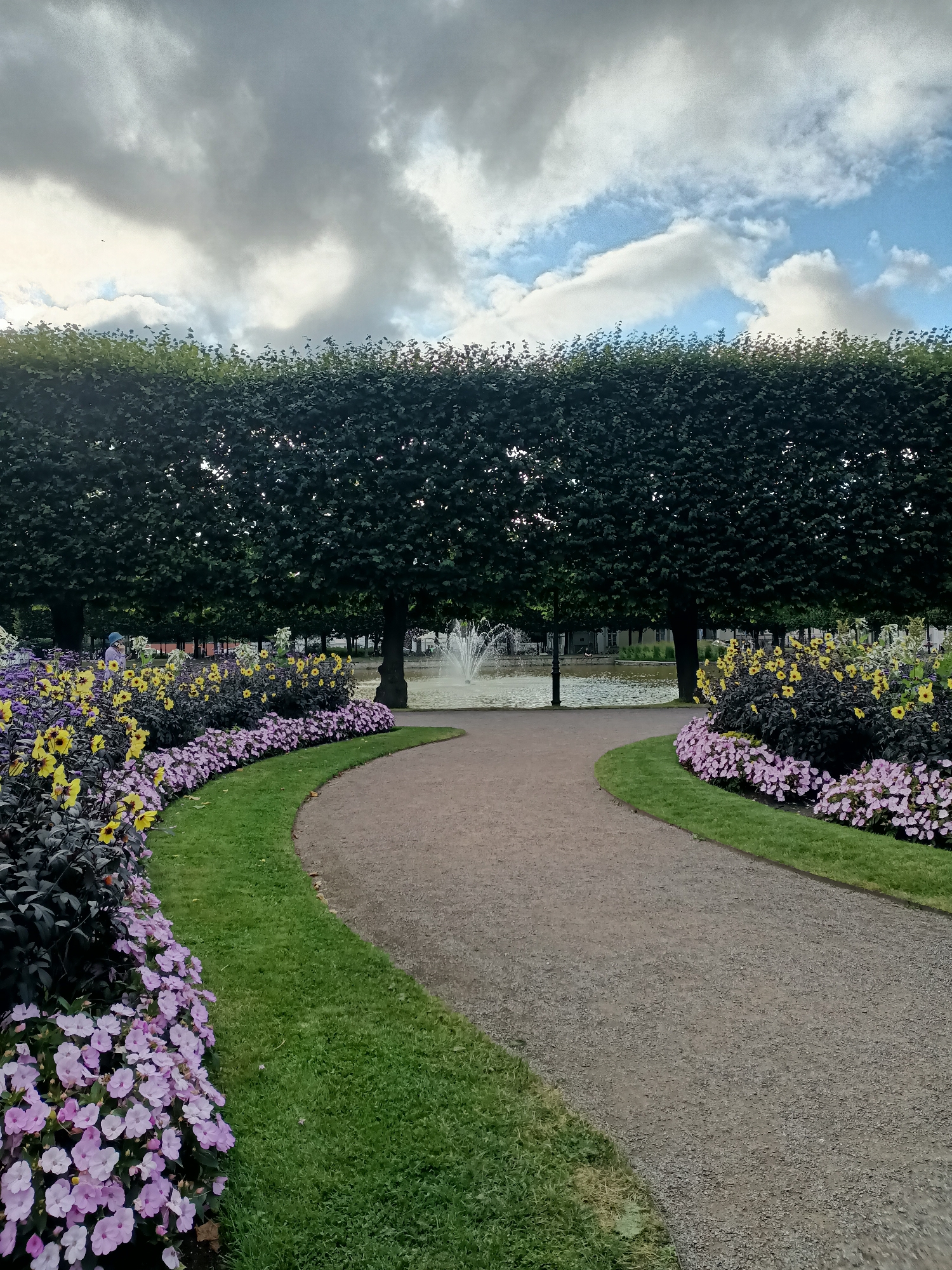 The image depicts a path in a park, curving to the right. On the left side of the path, there are flower beds with pink and yellow flowers, bordered by dark green foliage. Beyond the flowers, there is a row of trimmed, dark green trees. In the background, a fountain is active in a body of water, and further back, buildings are visible. The sky above is partly cloudy, with dramatic grey and white clouds against a blue sky. The scene is set in Tallinn, Estonia, specifically in Kadriorg Park.