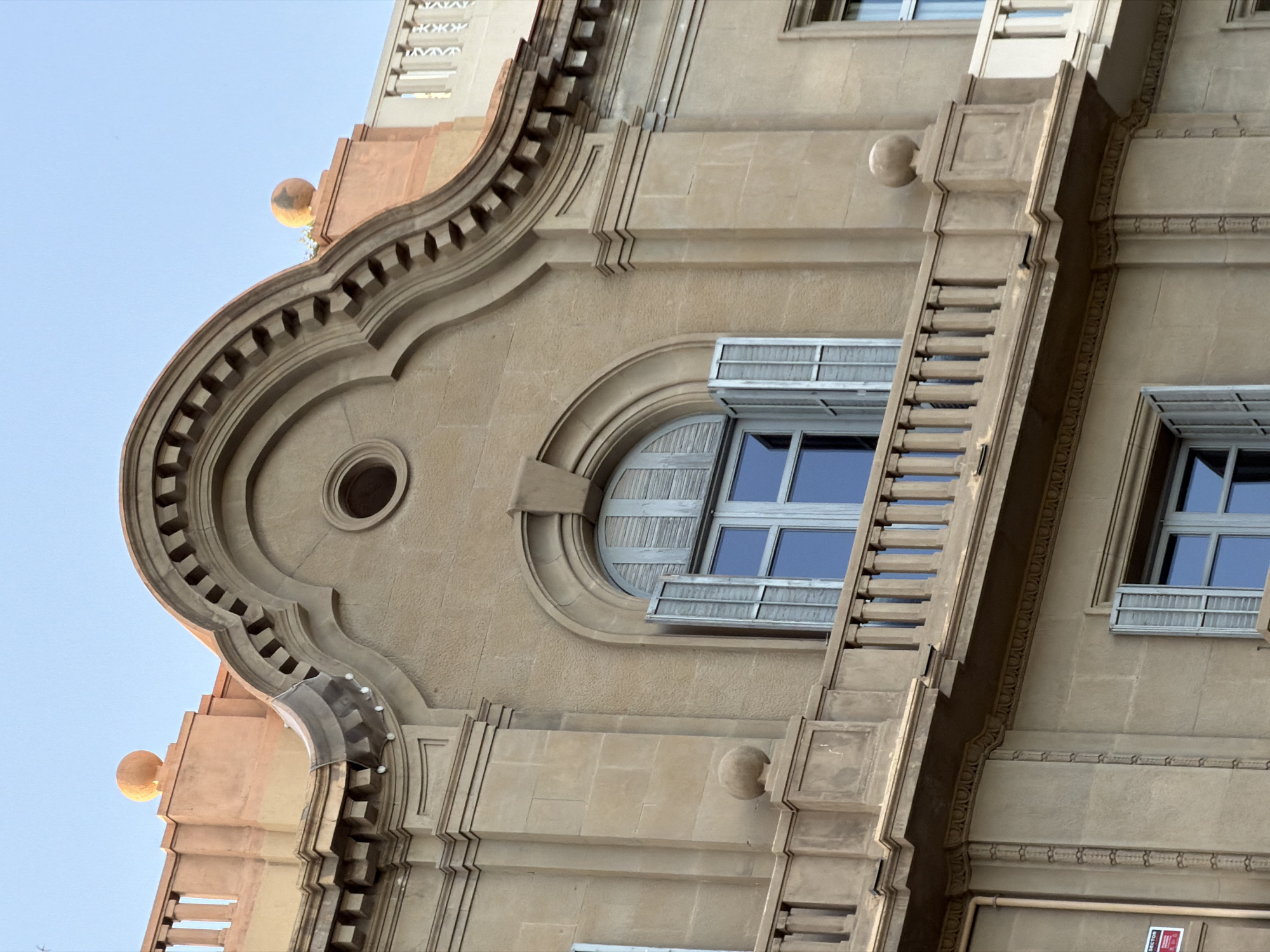 The image shows the facade of a building in Barcelona, Spain, with an arched window and shutters. The building's architecture is ornate, with decorative molding and a balustrade. The sky is clear and blue, suggesting it is daytime. The image is a close-up, focusing on the upper part of the building.