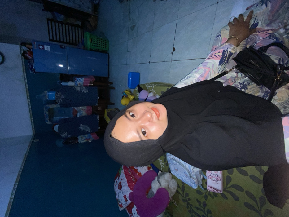 A person wearing a black hijab poses in a room. They are positioned on a bed with a green floral pattern and several plush toys. The person has fair skin, light brown eyes, and wears orange lipstick. An arm with a bracelet is visible next to a black purse. The room is dimly lit and contains furniture, including a blue cabinet and a storage rack with blue containers. The walls are blue and the floor appears tiled.