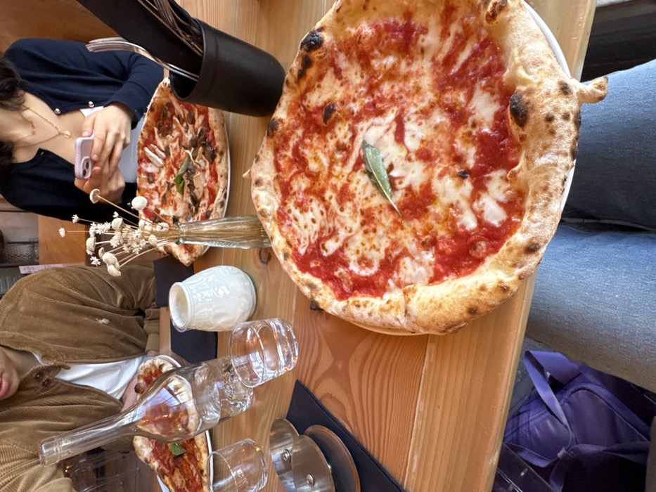 The image captures a top-down, slightly angled view of a dining table, rotated ninety degrees counter-clockwise. The scene shows two pizzas and several other items on a light wooden table, with parts of two individuals visible.
In the foreground, dominating the right side of the frame, is a large, round Neapolitan-style pizza. It features a prominent, puffed crust with visible charring, indicating a high-temperature bake. The pizza is topped with a vibrant red tomato sauce and melted white mozzarella cheese, which has a bubbly texture. A single green leaf, likely basil or sage, is placed near the center of the pizza.
Towards the middle and left side of the table, another smaller pizza is visible, partially obscured by other items. This pizza appears to have toppings like mushrooms and possibly other vegetables or herbs, differing from the simpler Margherita-style pizza in the foreground.
Several dining accessories are scattered across the table. A tall, black cylindrical holder filled with various cutlery (forks and knives) is positioned towards the top left. Below this, a slender glass vase holds a small bunch of dried white, spherical flowers or grass. A small, white, textured ceramic jug or vase is also on the table.
Numerous clear glass items are present: several empty drinking glasses are arranged, some upright and some tilted. An empty glass bottle, shaped like a wine bottle but possibly decorative, lies on its side, reflecting light. A silver pizza cutter with a black handle rests on a black placemat.
Two individuals are partially visible at the table. In the upper left corner, a person with dark hair is seen wearing a dark blue cardigan with light buttons over a white top. They are holding a light pink smartphone, seemingly taking a picture or video of the food. A delicate gold necklace with a circular pendant is visible around their neck. In the lower left, another person is partially shown, wearing a brown corduroy jacket over a white top. Only their torso and arm are visible, and they appear to be looking down at the table.
To the far right of the image, the leg of a person wearing blue jeans is visible, suggesting someone is seated at the table on a bench or chair. In the very bottom right corner, a purple bag, possibly a backpack or tote, is partially visible on the floor or a nearby seat.
The overall impression is that of a casual meal at a restaurant or cafe, with people enjoying food and one person capturing the moment with their phone. The lighting suggests daytime.