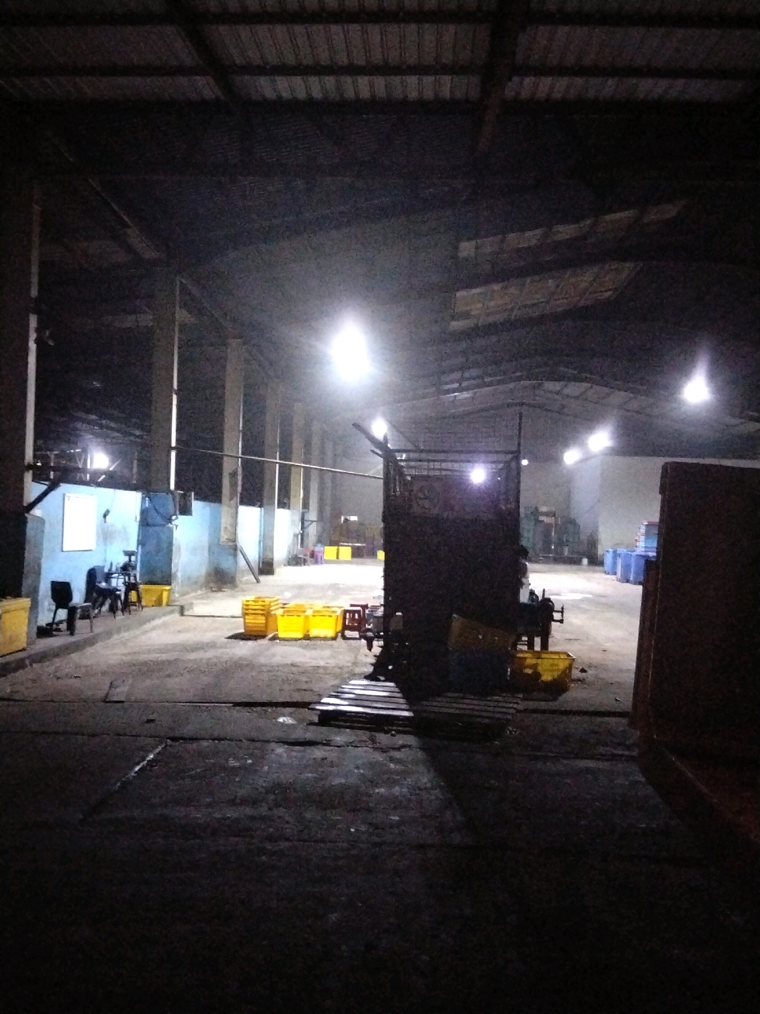 The image depicts the interior of a dimly lit warehouse or industrial building, likely in Moro, Indonesia. The structure has a high, corrugated metal roof supported by numerous concrete pillars. Artificial lighting is sparse, with several bright, white lights illuminating sections of the space. The floor is concrete, and appears dirty and uneven.
In the foreground, the concrete floor is dark and textured, with visible cracks and patches. A few wooden pallets are stacked on the ground. Further into the building, a large, dark, metal structure, possibly a piece of machinery or a vehicle, is centrally located. To the left of this structure, there are several bright yellow plastic bins, some stacked.
Along the back wall on the left side of the image, there is a blue-painted partition with a window and a closed door. Several chairs and small tables are arranged near this partition, along with more yellow bins. The wall is rough and appears to be made of concrete blocks.
The mid-ground of the warehouse shows more yellow bins and some red stools. In the distance, the warehouse extends further back, with more pillars and what appear to be stacks of blue barrels. The overall impression is of a functional, but somewhat disused or unkempt, industrial space. There are no people clearly visible in the image. The lighting creates strong contrasts and deep shadows, contributing to a somewhat eerie atmosphere.