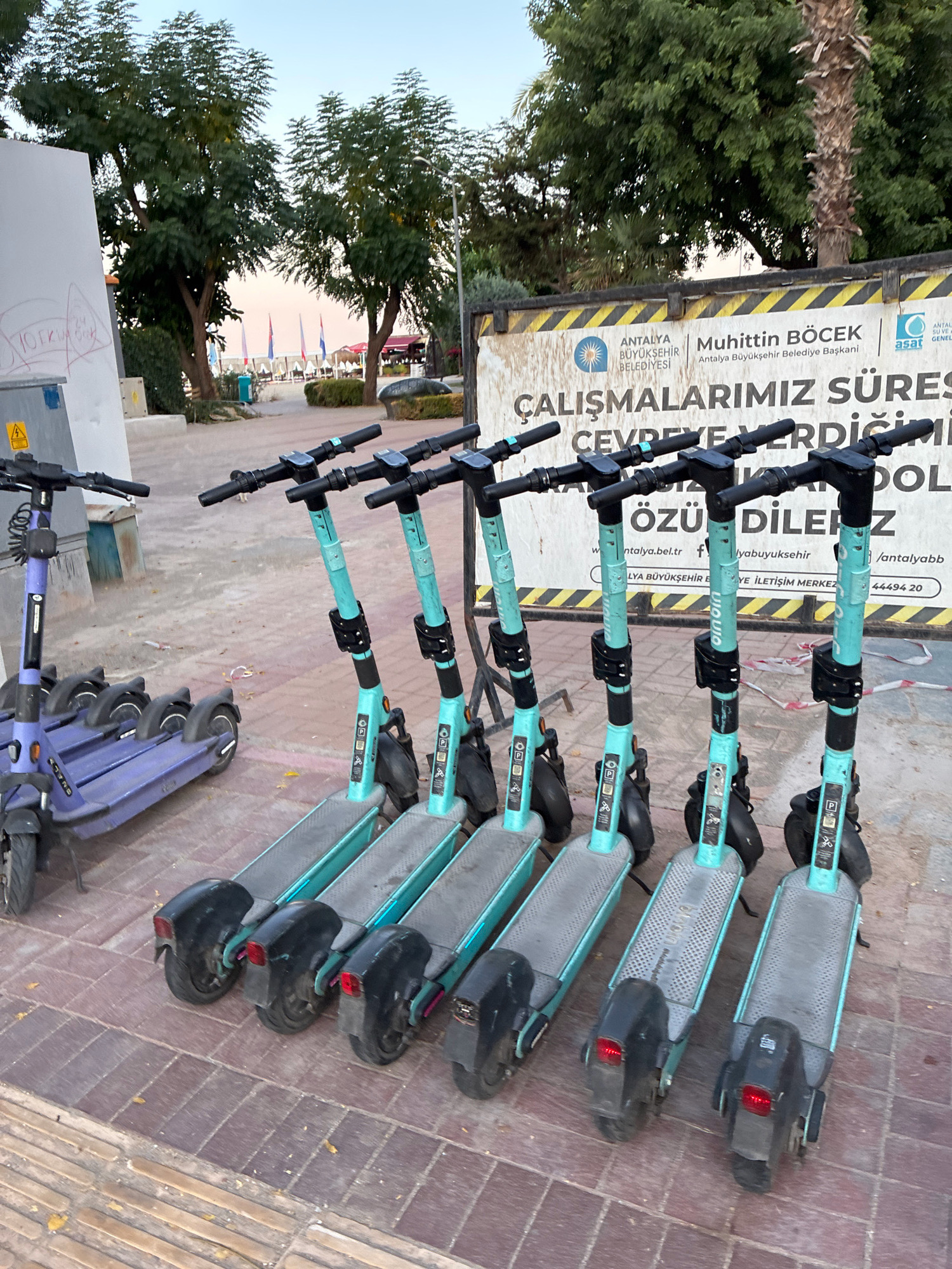 This image shows a row of parked electric scooters on a sidewalk in Alanya, Turkey. The scooters are predominantly teal with black handlebars and are lined up in front of a construction banner. The banner features Turkish text and logos, indicating a public works project by the Antalya Metropolitan Municipality. In the background, trees and a glimpse of a beach with flags suggest a coastal urban setting. A purple scooter is visible to the left, parked separately. The sidewalk is paved with reddish-brown bricks. The lighting suggests it is either early morning or late afternoon. There are no people or animals clearly visible in the frame.