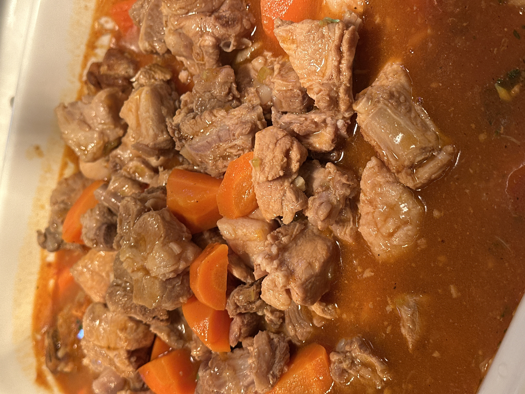This image depicts a close-up, overhead view of a cooked stew or braise served in a white, rectangular dish. The scene is located in Púng Luông, Vietnam. The dish primarily contains numerous irregular chunks of light brown meat, consistent with pork, interspersed with bright orange, uniformly diced carrots. All ingredients are coated and partially submerged in a thick, reddish-orange sauce that exhibits a glossy surface. Small, indistinct flecks, possibly herbs, spices, or finely minced vegetables, are visible within the sauce. The meat pieces show varying textures, with some appearing lean and others presenting visible fatty or connective tissue. The carrots appear softened from cooking. The white serving dish has slightly angled edges. The background outside the immediate dish is out of focus and indistinct. No people, specific actions, or external environmental details beyond the serving dish are discernible.