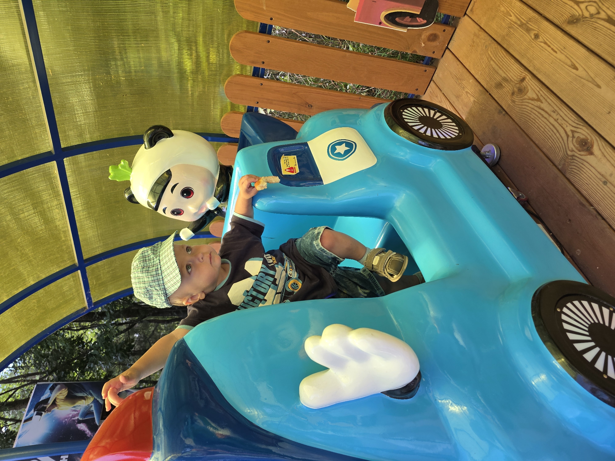 A young Caucasian boy wearing a plaid baseball cap, a dark t-shirt with a graphic of a car and the word "VICTORY," and denim shorts sits in a bright blue toy car. He is holding a small piece of bread in his right hand, near his mouth. To his right, a cartoonish panda figurine is visible. The toy car appears to be part of an amusement park ride, set up on a wooden platform. In the background, a yellow translucent canopy is supported by blue beams, and a wooden fence separates the ride area from vegetation and trees. A poster featuring a person wearing a VR headset is visible in the upper left corner. The overall scene suggests an outdoor recreational setting, likely an amusement park or playground. There is no information in the image to suggest the location is the city of Chaikovsky, Russia.